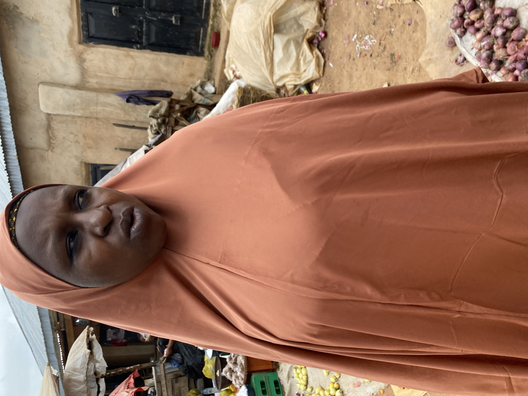 A woman in a brown hijab stands in what appears to be an outdoor market in Bukuru, Nigeria. The woman's face is visible, looking to her right with her mouth slightly open. Her hijab covers her hair and neck, and a portion of her upper body is visible through the fabric. In the background, there are market stalls with various goods, including produce like onions and what appears to be fruit, as well as sacks and other items. A building with a corrugated metal roof and a wooden door is also visible. The ground is dirt.