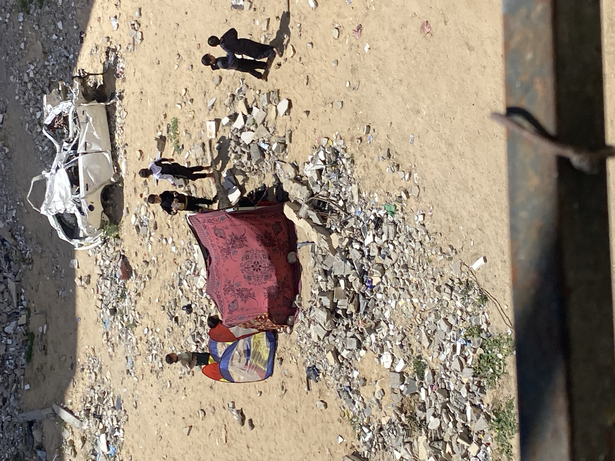 This image depicts a scene in what appears to be the aftermath of destruction, likely in Khan Yunis, Palestinian Territory. A severely damaged white vehicle is visible on the left side of the image. The ground is covered with rubble, debris, and some sand. There are groups of people scattered throughout the scene: Two individuals in darker clothing are standing together towards the top, while another two individuals are located near the wrecked car. Further down, under a patterned blanket, two other individuals are present; one of them is lying down under a colorful blanket. The entire scene is captured from an elevated perspective, possibly from a nearby building.