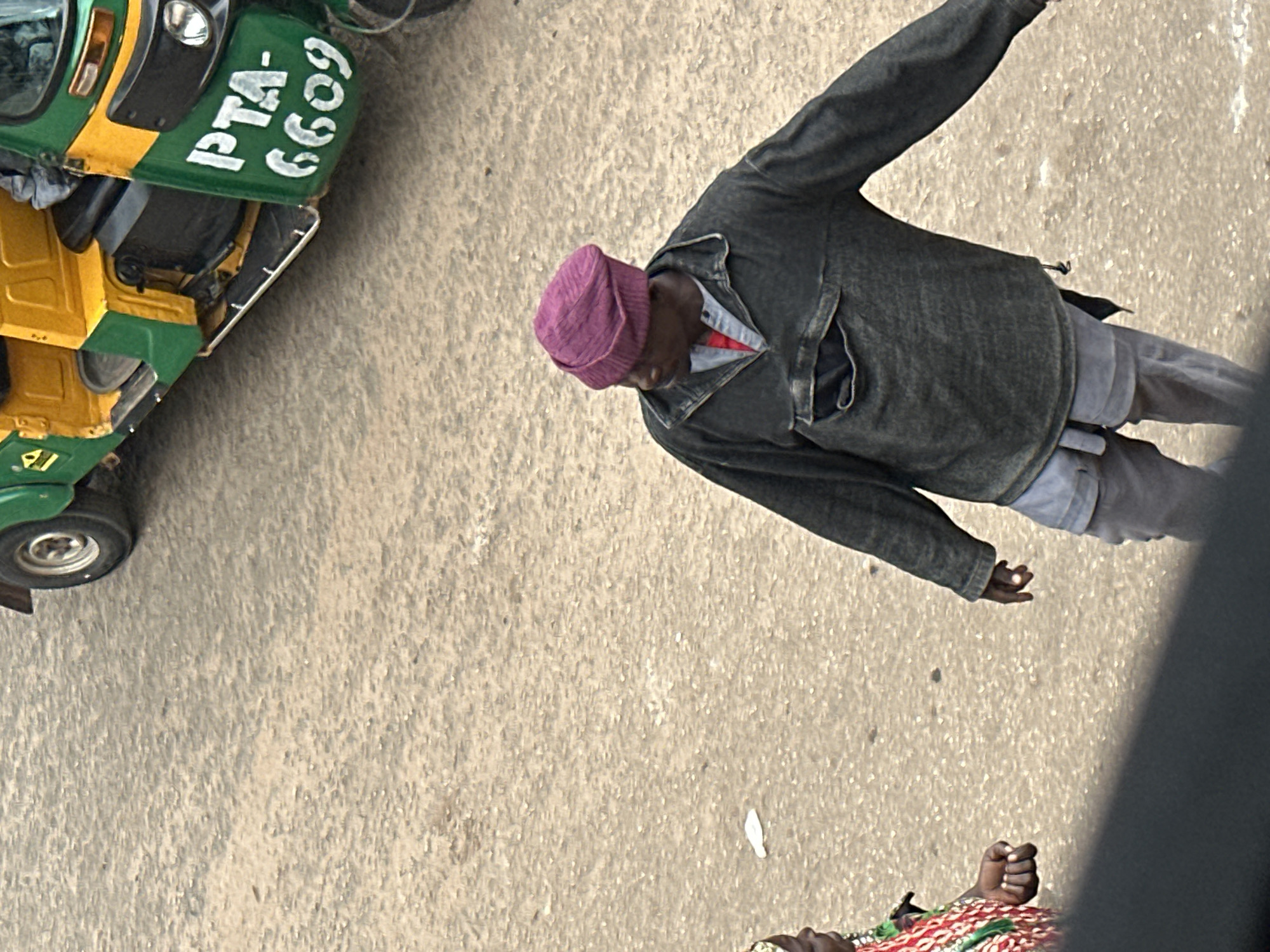 An overhead shot captures a street scene in Bukuru, Nigeria. A green and yellow auto-rickshaw, identified by the license plate "PTA-6609", is parked on a dusty, tan-colored road. Two individuals are visible. In the foreground, a man wearing a dark, long-sleeved garment over a red undershirt and light gray shorts, topped with a purple beanie, stands looking towards the left. To his left, partially visible from the shoulder down, is a person in a vibrantly colored garment. The ground is characterized by its rough, uneven texture.