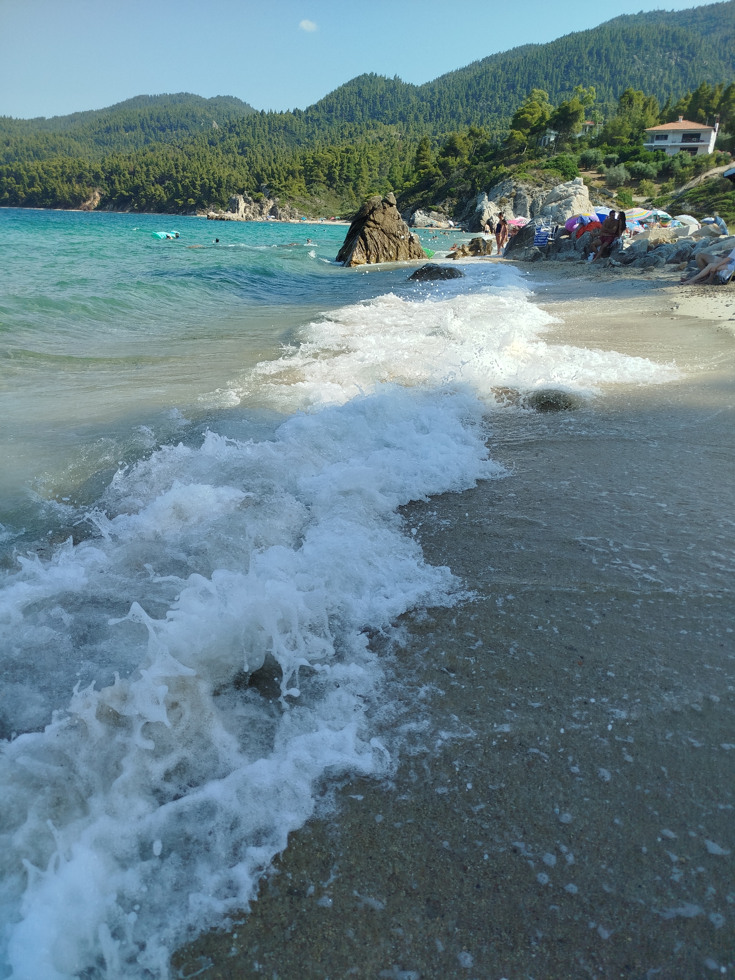 The image depicts a sunny day at a beach in the Sithonia Municipal Unit, Greece. The foreground is dominated by a breaking wave, with white foam and spray crashing onto the sandy shore. The water is a clear turquoise color, transitioning to a deeper blue further out. In the background, a lush, green, pine-covered hillside rises steeply from the coastline. Several rocks, some large and dark, are scattered along the shore and in the water.
Further back along the beach, a small group of people are visible. Some are seated under colorful beach umbrellas, while others appear to be walking or wading in the shallow water. Beach chairs and towels are spread out on the sand. A few buildings, possibly houses, are nestled into the hillside amidst the trees. The sky is a clear, bright blue with no clouds. The overall atmosphere is one of a serene and picturesque beach vacation.