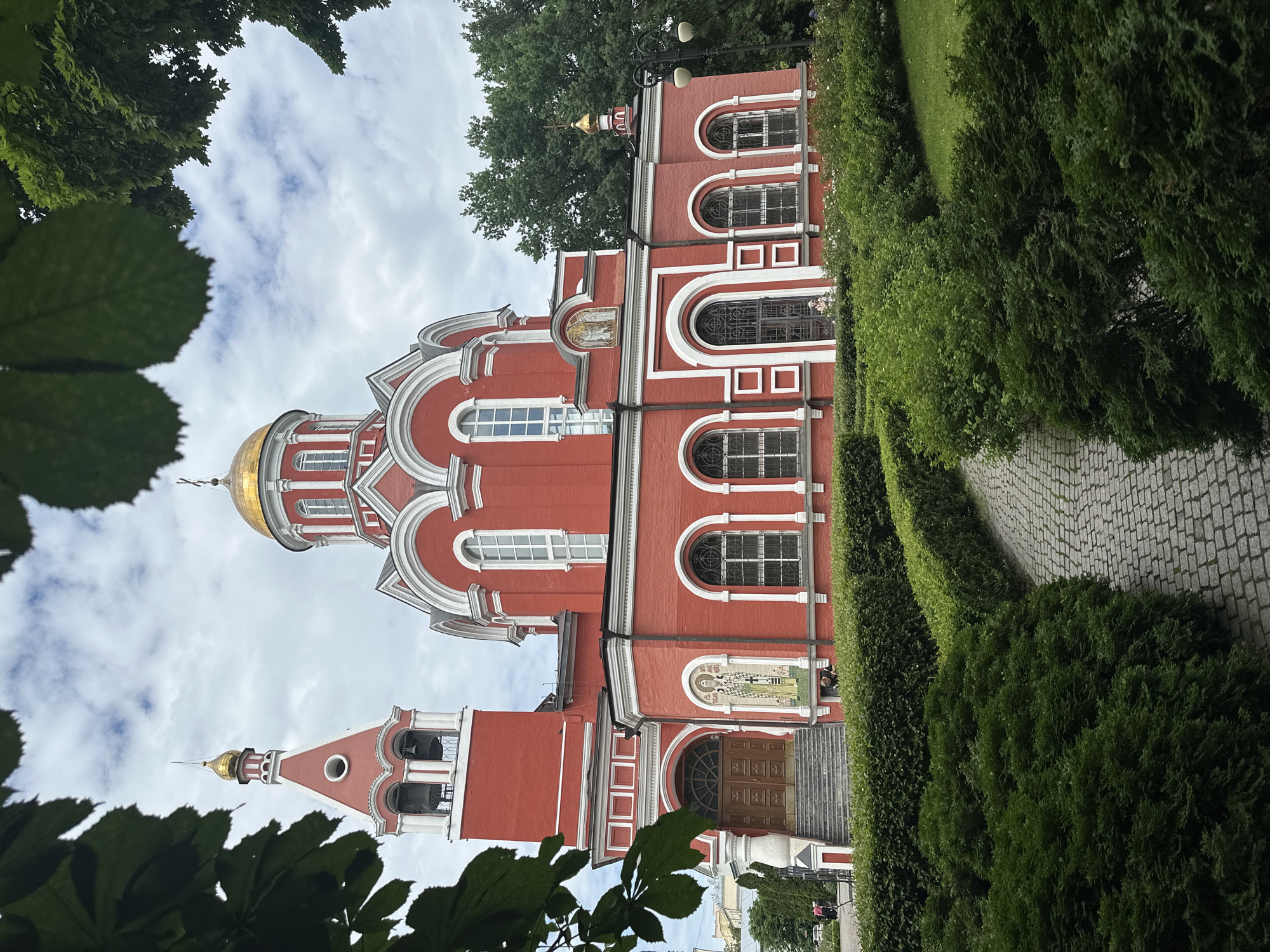 The image shows a red orthodox church with white trim and gold domes, partially framed by green foliage in the foreground. A paved walkway with cobblestones winds through manicured hedges leading towards the church's entrance, which is accessed by a set of stairs. The church has a bell tower on the left and a main dome in the center. Mosaics adorn the exterior walls. The sky is partly cloudy. Several people are visible in the distance near the church entrance and on the grounds. Trees and more greenery surround the building. The scene appears to be an exterior view of a religious site on a day with overcast skies.