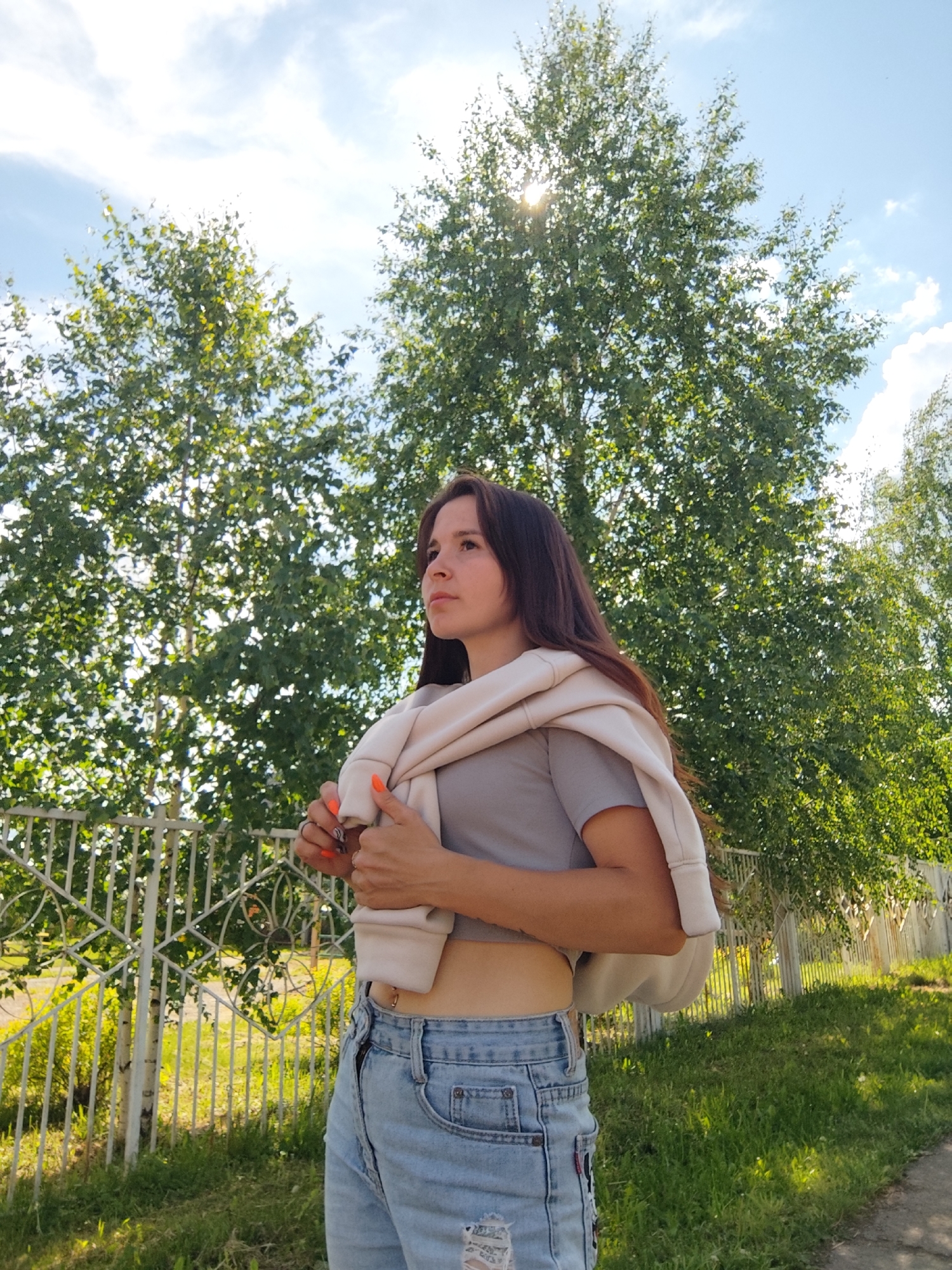 A young woman stands outdoors on a sunny day. She has long brown hair and is wearing a gray crop top and blue jeans. A beige sweatshirt is draped over her shoulders. Her nails are painted orange. The woman is standing next to a white metal fence. Behind her are green trees, with sunlight filtering through the leaves. The location is Фоки, Russia.
