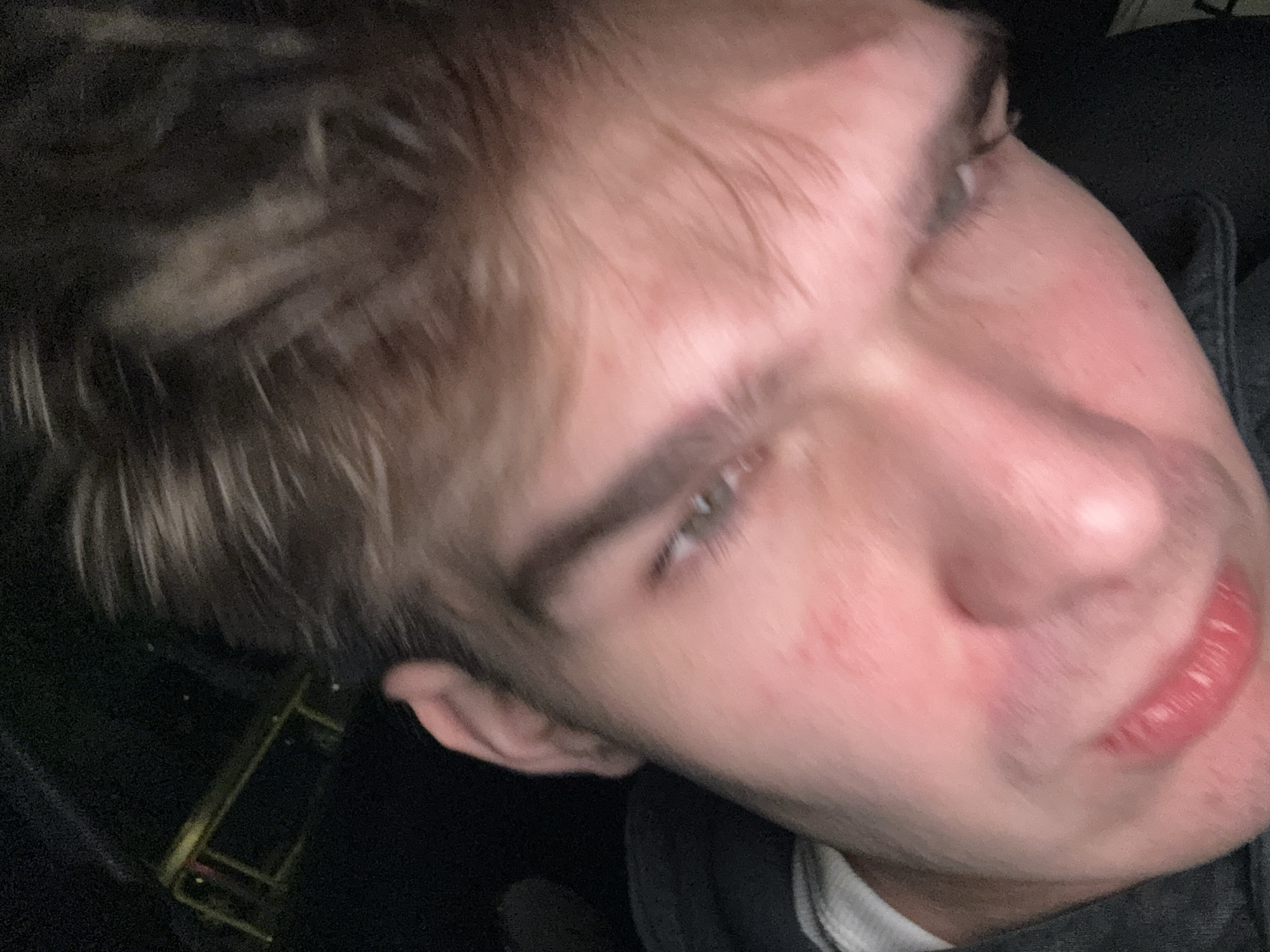 The image is a close-up, blurred, and low-angle selfie of a person. The person's face is partially obscured by hair. The lighting suggests an indoor environment with dim ambient light, possibly from a car interior or a dimly lit room. The background is dark and indistinct, with a hint of a metallic structure with yellow accents visible on the left side, which is too blurry to identify specifically but might suggest a stage prop or equipment. The overall impression is one of casual, candid photography. There is no discernible location in Warsaw, Poland, indicated by the visual elements of the image.