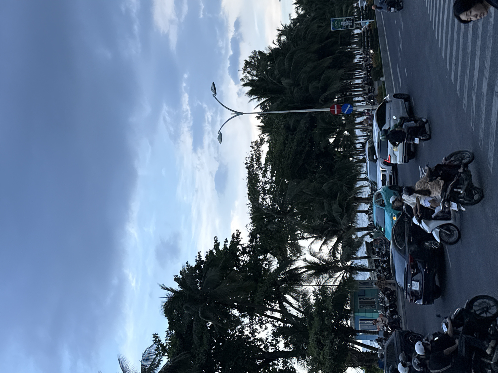 This image captures a busy street scene in Nha Trang Ward, Vietnam, near the coast, as indicated by the presence of palm trees and a glimpse of the ocean. The sky is overcast with a mix of blue and gray clouds, suggesting either early morning or late afternoon.
The foreground shows a road with several vehicles, predominantly motorcycles, and a few cars. People are riding motorcycles, some wearing helmets, and others are walking. There's a crosswalk in the lower right portion of the image.
In the middle ground, the road continues with more traffic, including a turquoise car, a black sedan with a license plate that appears to read "29A 150.89," and a white SUV. There are also many parked motorcycles along the side of the road. A distinctive teal building with a blue roof stands near the water's edge, possibly a small shop or a rest stop.
The background is dominated by dense foliage, primarily palm trees and other lush tropical vegetation, lining the street and extending towards the ocean. A street lamp with two lights is prominently visible on a pole in the center of the frame. Several traffic signs, including a red and white circular sign and a blue circular sign, are also present. The overall impression is of a vibrant, active urban environment integrated with a coastal setting.