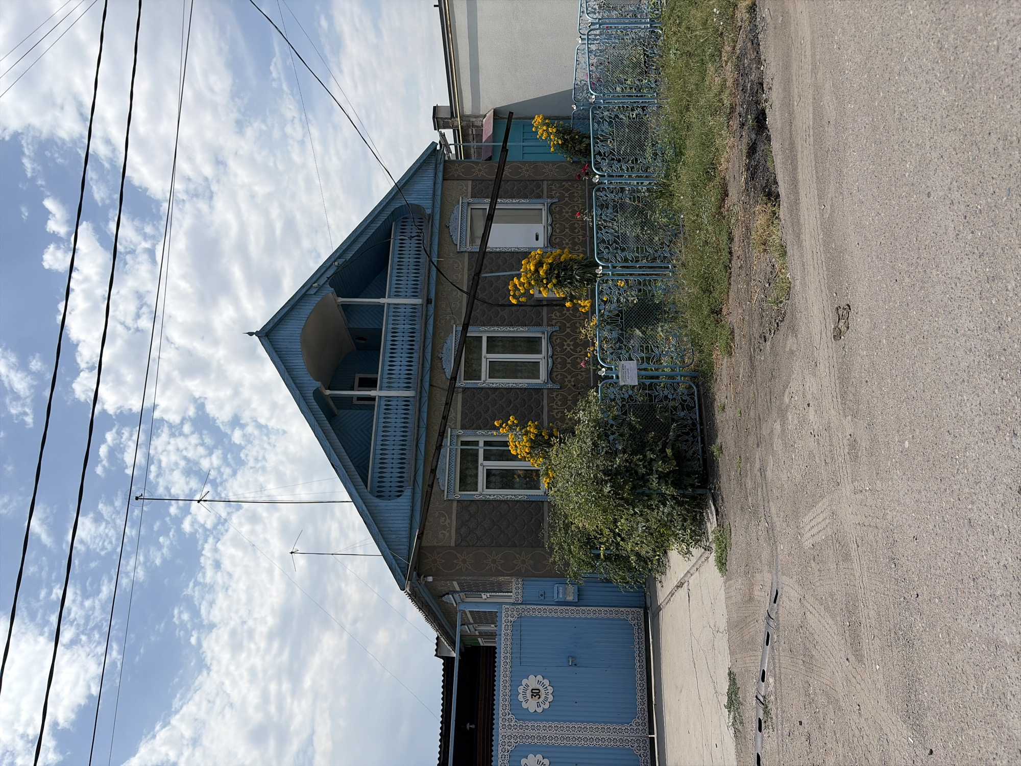 This is a front-facing, eye-level photograph of a house in Novopokrovka, Kyrgyzstan. The house is a two-story structure with a blue-painted, gable-style roof and a balcony on the second story. The exterior walls of the house are gray with a textured pattern. The windows on the first floor are adorned with blue decorative trim. A blue metal gate with ornate white scrolling leads to the property, featuring the number "30" on a circular plaque. The yard is enclosed by a blue, decorative metal fence. In front of the house, there are several bushes and a cluster of bright yellow flowers. A mailbox is attached to the front gate. Overhead, power lines crisscross the sky, which is partly cloudy. The ground in front of the gate is paved, transitioning to a gravel road with tire tracks. To the right of the house, a section of a white wall is visible. The overall scene depicts a residential property on a sunny day.