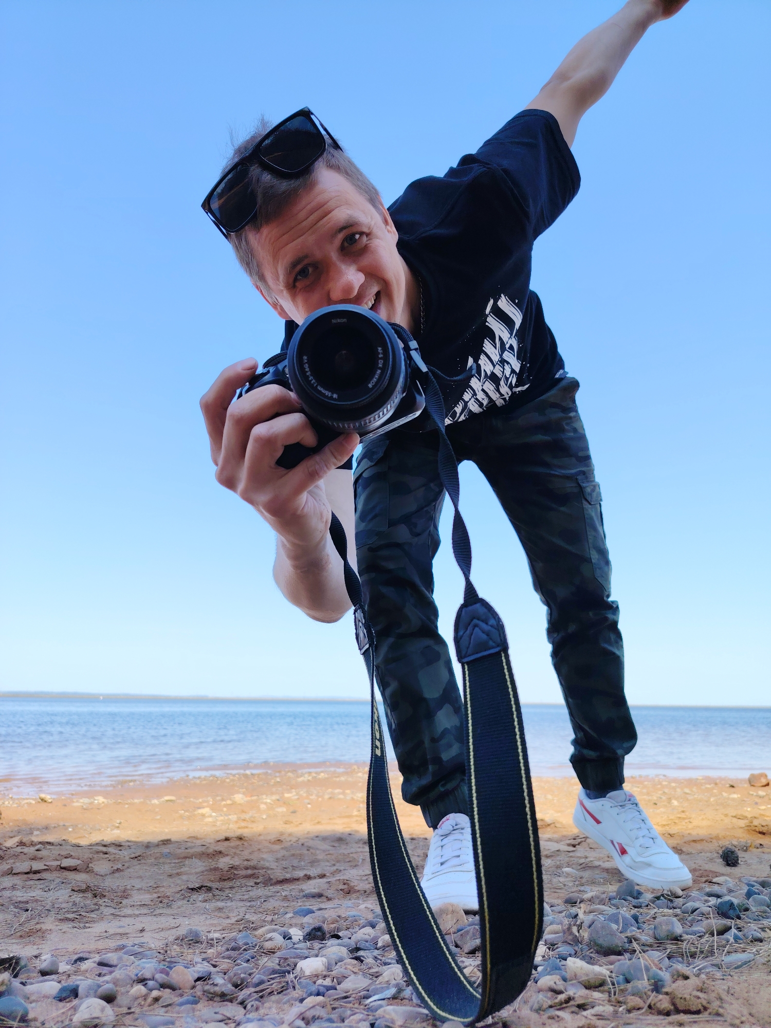 A man is posing at a beach on the Kama River in Chaikovsky, Russia, holding a camera pointed at the viewer. He has short brown hair, sunglasses perched on his head, and is wearing a black t-shirt with white print, camo pants, and white sneakers. He is smiling and extending his right arm upwards. The camera is a DSLR with a black strap. The beach is sandy with pebbles and small rocks. In the background, the Kama River is visible under a clear blue sky.
