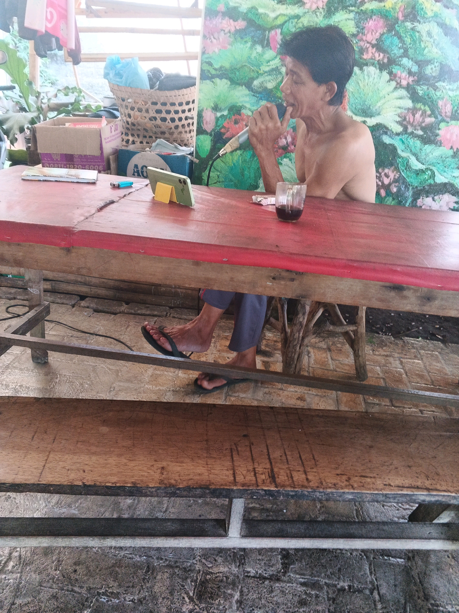The image depicts a man sitting at a red wooden table with a microphone in his hand, positioned near his mouth as if singing or speaking. He is shirtless and wearing dark pants and flip-flops. On the table, there is a glass containing a dark liquid, a lighter, a phone propped up on a stand, and some papers. Behind the man, a colorful mural with floral motifs is visible. The setting appears to be an outdoor or semi-outdoor area, with wooden structures and what looks like a tiled floor. The overall impression is that of a casual, perhaps recreational, moment. The image was taken in Ungaran, Indonesia.