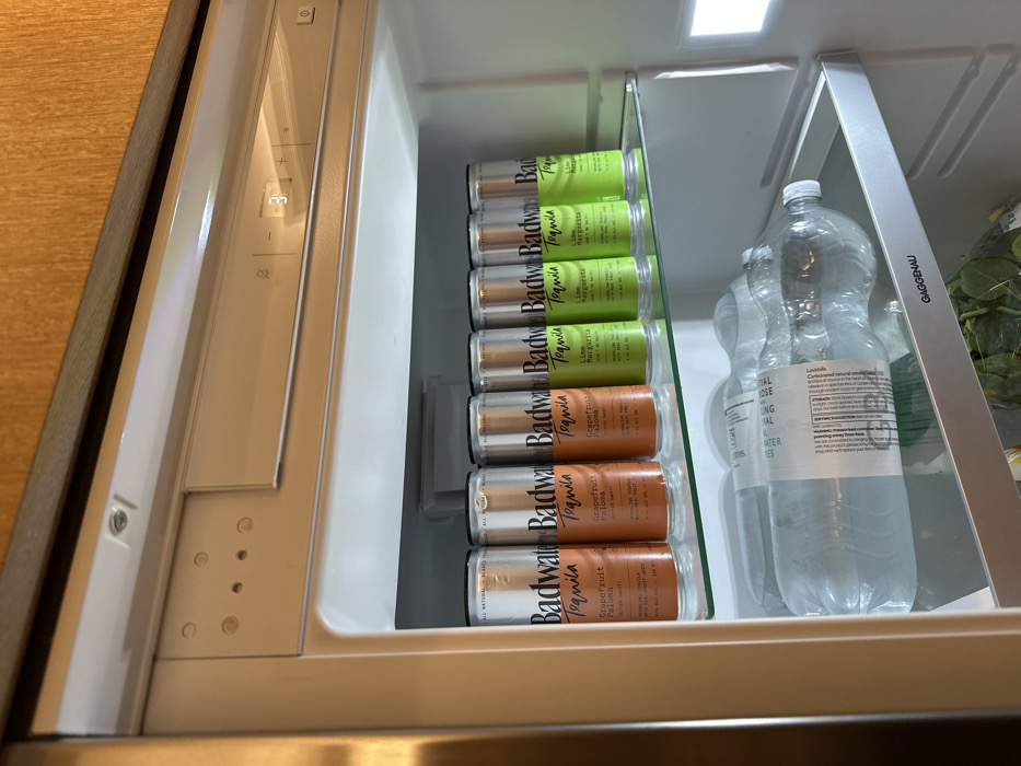 An open refrigerator shelf contains a neat row of "Badwater Tequila" canned cocktails. Three cans feature a brown "Grapefruit Paloma" label, followed by three green cans labeled "Lime Margarita". Behind the canned cocktails are two large clear plastic bottles of sparkling water. A container of fresh spinach sits in the upper right corner. The refrigerator's temperature control panel is visible on the left side of the image.