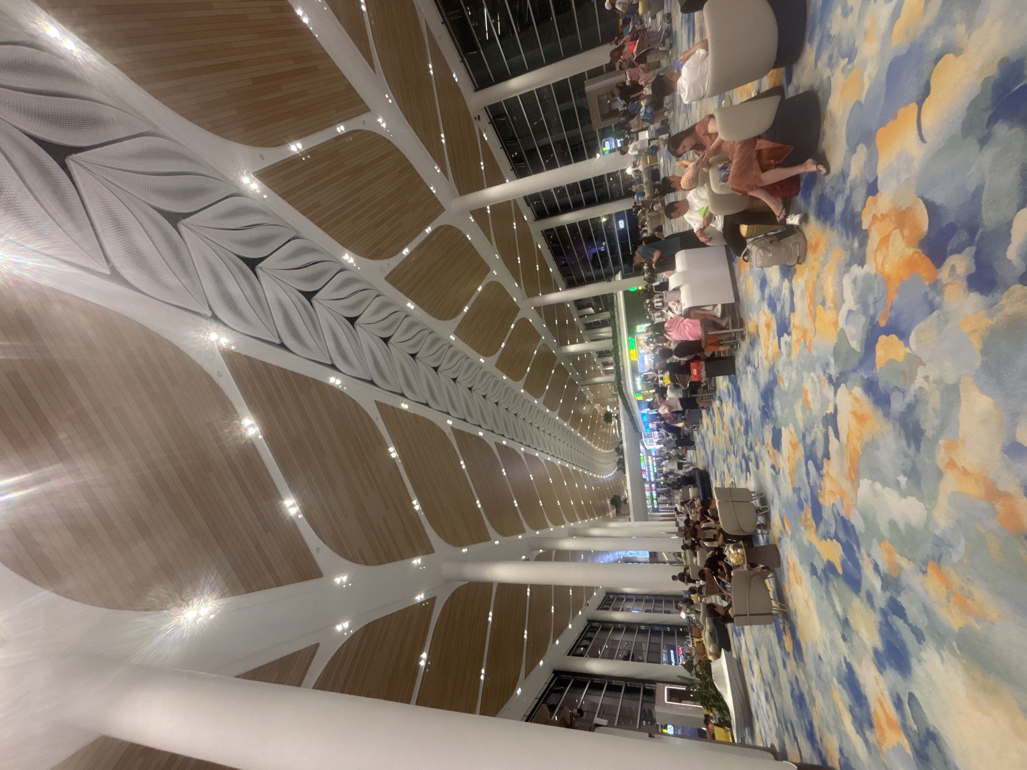 The image depicts a modern airport terminal waiting area in Baiyun District, China. The ceiling features a striking architectural design with white, organic-shaped beams resembling leaves against a backdrop of wooden panels. Numerous spotlights are embedded in the ceiling. Large white pillars support the structure. The floor is covered with a vibrant, abstract carpet featuring patterns of blue, orange, and white. People are seated in a variety of modern, upholstered chairs and sofas arranged in small groups and individually. Many individuals are looking at their phones or conversing. Some are seen with luggage. Shops and information displays are visible in the background through large glass windows and open spaces. The overall impression is one of a spacious, well-lit, and contemporary public space designed for travel.