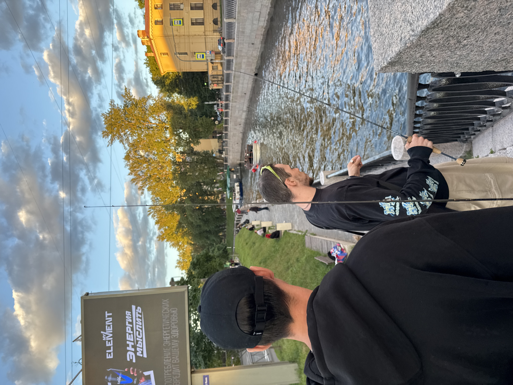 The image depicts two men fishing on the embankment of the Griboyedov Canal in Saint Petersburg, Russia, during the late afternoon or early evening, evidenced by the golden light illuminating the buildings and trees. The man in the foreground, facing away from the camera, is wearing a black hooded sweatshirt and a black baseball cap. The second man, partially visible and facing the canal, is wearing a black t-shirt with graphics on the sleeves and light-colored shorts. Both men are holding fishing rods. Several other people are visible along the canal bank, some sitting on benches and others standing. A boat is moving on the canal. A large billboard advertising "MY ELEMENT" energy drinks is situated to the left of the frame. The canal's water reflects the sky and the surrounding trees. The architecture of the buildings along the canal is characteristic of Saint Petersburg, with warm yellow facades and classical detailing. The sky is partly cloudy, with the clouds illuminated by the setting sun.
