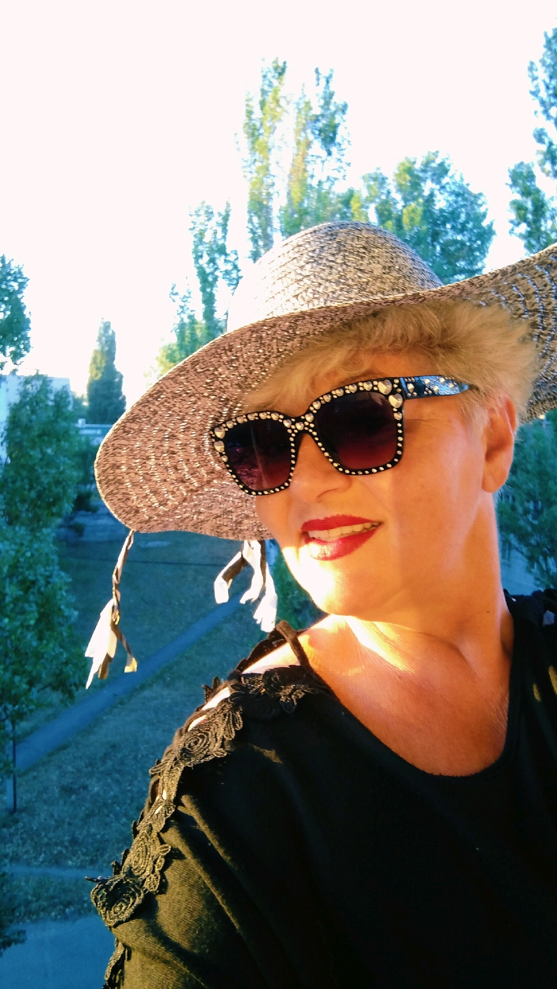 A close-up, frontal selfie captures a woman wearing a wide-brimmed, woven straw hat adorned with decorative elements, and oversized, sparkly sunglasses. The woman has short, light-colored hair, and her lips are painted red. She is wearing a black top with lace detailing on the shoulders and straps. The background appears to be an outdoor setting with trees and a sky illuminated by sunlight, suggesting a daytime shot. The overall impression is that of a woman posing for a casual, outdoor photograph. The specific location in the city of Horishni Plavni, Ukraine, is not discernible from the image itself.