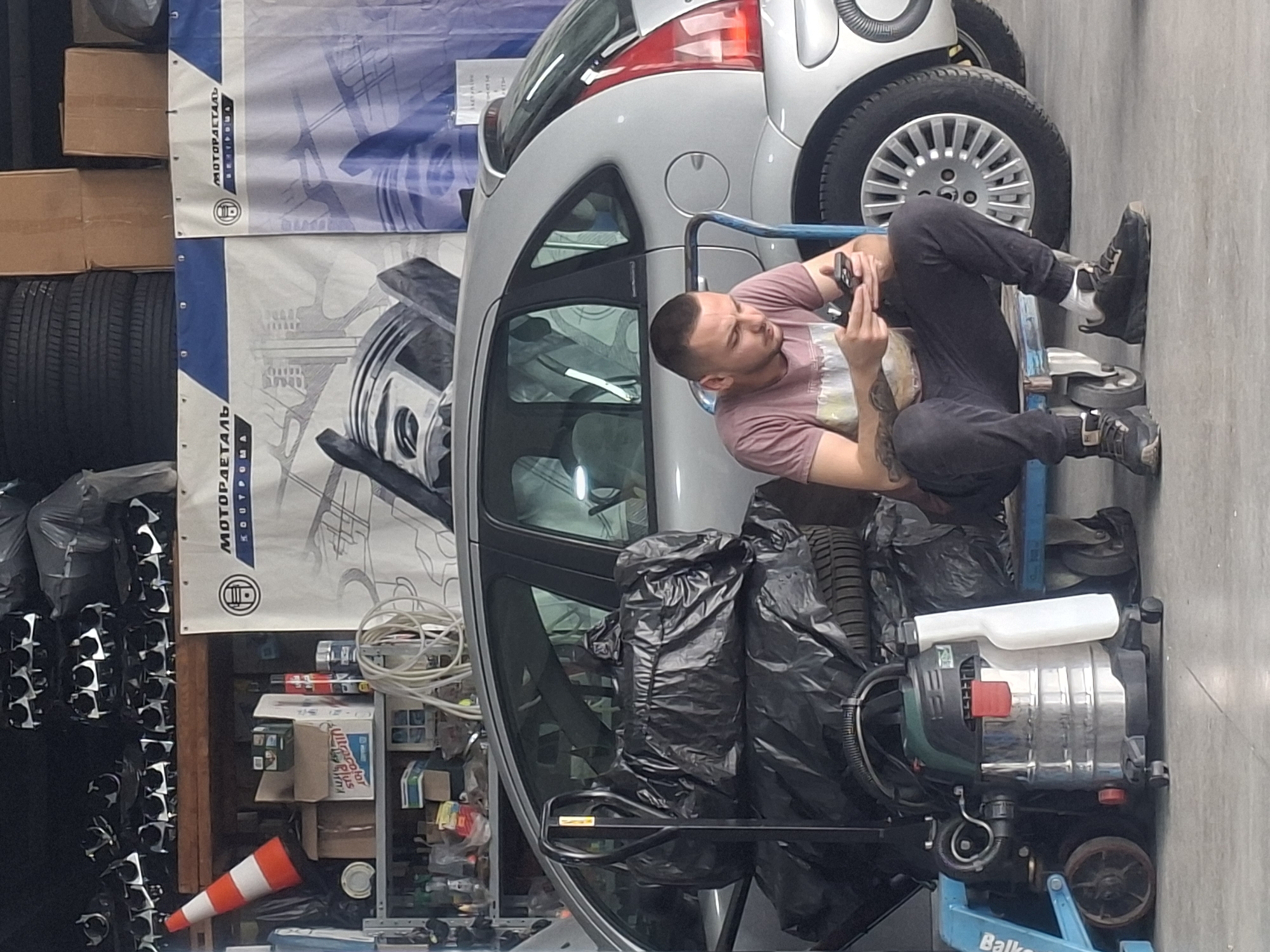 The image depicts a man sitting on a blue wheeled cart in what appears to be a garage or workshop setting in Unknown, Unknown. The man is holding a mobile phone, looking at the screen. He is wearing a pink t-shirt and black pants. A silver car is parked nearby. A pressure washer is on the cart with the man. Bags of tires are nearby as well. A tire rack and automotive supplies are visible in the background. Automotive-related banners are hanging.