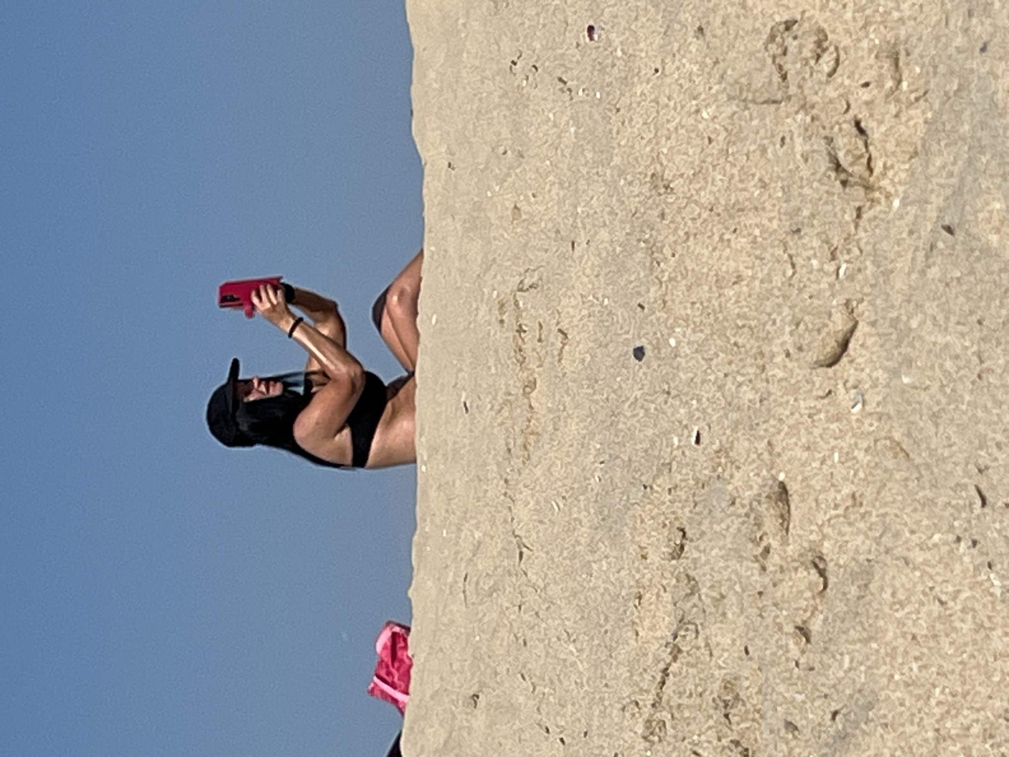 A woman in a black bikini and a black baseball cap is sitting on a sand dune, holding a pink smartphone. She is looking at the phone and appears to be taking a picture. The background is a clear blue sky. To the left, a pink bag is visible on the sand dune. The image is taken on a bright, sunny day. The location is the beach in Nesebar, Bulgaria.