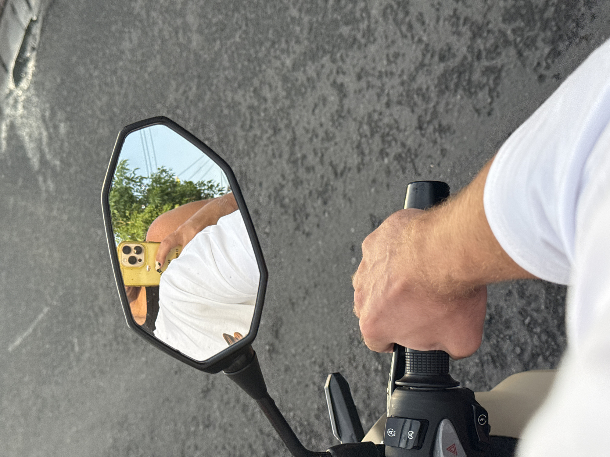 The image captures a first-person perspective from a motorcycle or scooter, looking down at the handlebar and side mirror, with a grey asphalt road as the background.
The main subject is the unseen rider, whose right arm and hand are visible, gripping a black, ribbed handlebar. The rider is wearing a white t-shirt. On the scooter's controls, several buttons are visible: one marked "OFF" with a left-pointing arrow, an "A" in a circle, another symbol resembling waves or "W" in a circle, and a prominent red hazard light triangle.
The left rear-view mirror reflects the person taking the photograph. In the mirror, a yellow smartphone with multiple cameras (suggesting an iPhone) is clearly visible in the person's hand, aimed at the mirror to capture the shot. The reflection also shows part of the person's arm and torso, consistent with wearing a white t-shirt. Beyond the reflection of the rider, the mirror displays a clear blue sky and lush green trees and foliage, indicating an outdoor setting.
The setting is an outdoor paved area, likely a road or street, characterized by its grey, textured asphalt surface. The lighting suggests it is daytime, with bright conditions and a clear sky, as seen in the mirror's reflection. The overall scene implies the scooter is either stationary or moving slowly in North Kuta, Indonesia.