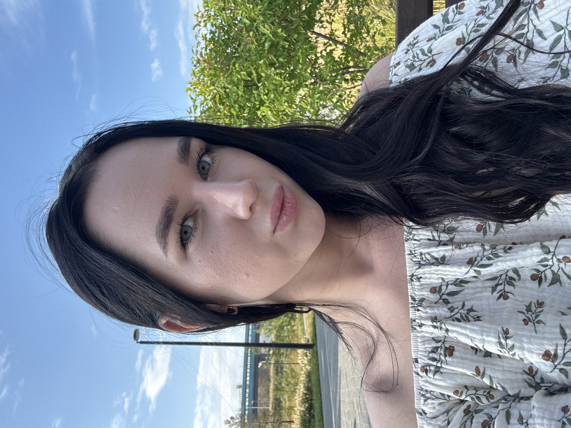 A young woman with long, dark hair and light eyes is captured in a selfie outdoors. She is wearing an off-the-shoulder white top with a floral pattern. Her face is turned slightly towards the camera, and she has a playful expression with her lips pursed. The background shows a clear blue sky with some clouds, a green bush, a pathway, and a street lamp. In the distance, there's a glimpse of a modern building with blue accents, possibly part of the infrastructure in Novosibirsk, Russia.
