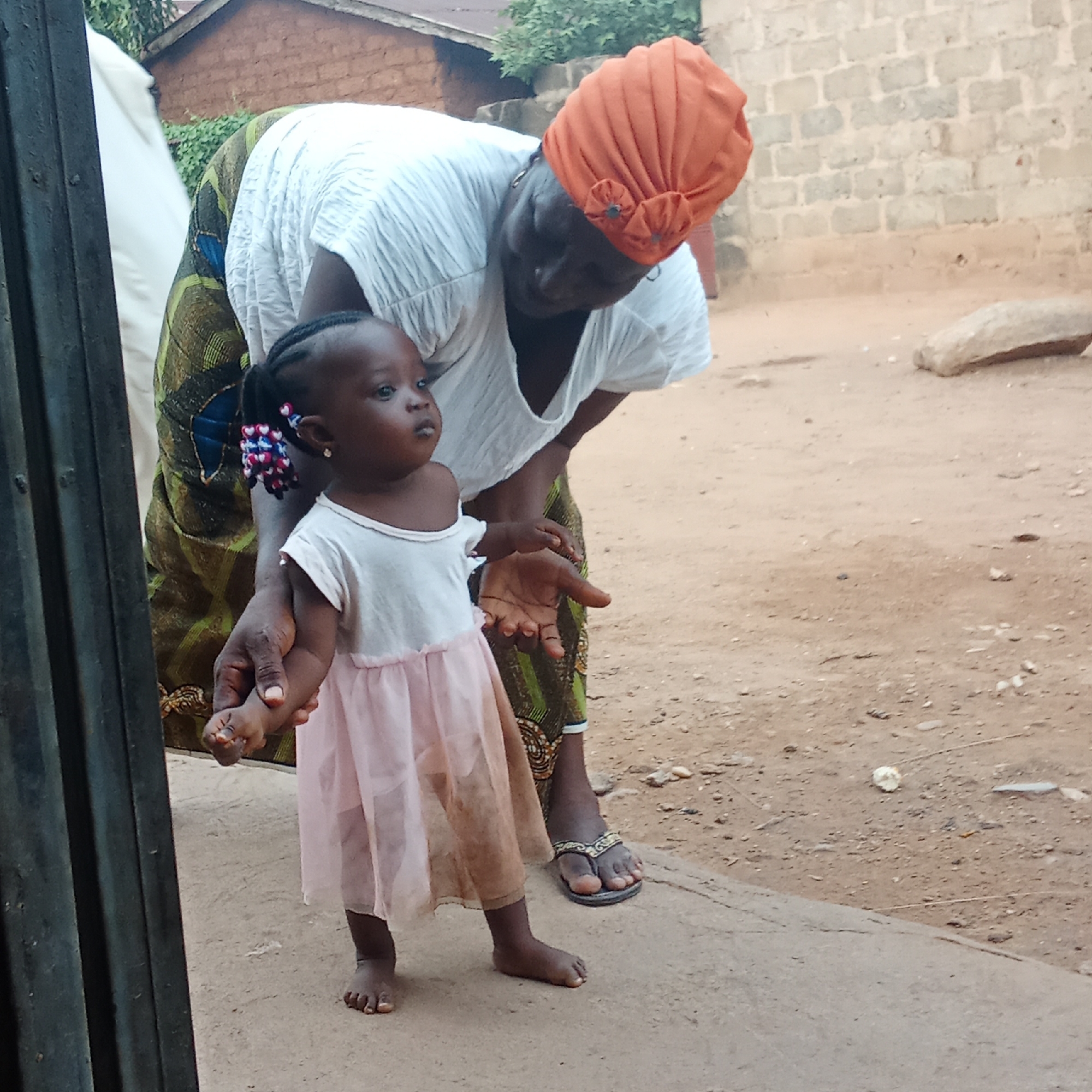 An eye-level medium shot shows an African woman and girl in an outdoor environment in an unknown city. The woman, who is bent over, has an orange headdress, a white short-sleeved shirt, and a patterned long skirt. The girl stands next to her with a white top and a light pink dress. The girl has colorful hair beads and is looking off to the side. The woman is holding the girl’s hand with her left hand and has her right hand extended. They are standing on a concrete surface, and behind them is a dirt ground with a wall. In the background, there is also greenery and a building with a brown roof. A dark, vertical object is on the left of the frame.