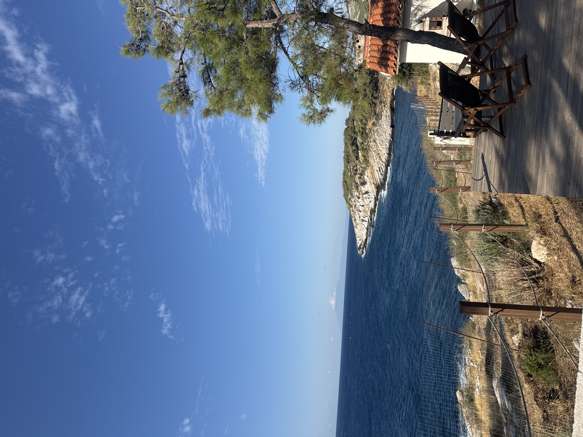 A view of the Aegean Sea from a wooden deck, featuring two empty lounge chairs, a small white building with a red tile roof, and a pine tree. In the distance, a rocky coastline stretches out, and a hazy mountain looms on the horizon. The sky is a clear blue with a few scattered white clouds. The scene appears to be in Greece.