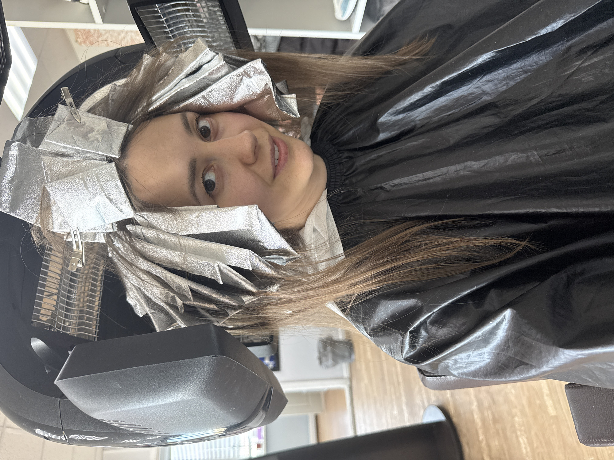A woman is seated in a black salon cape with silver foils in her hair, presumably undergoing a hair coloring treatment. She is positioned under a salon hair dryer. The scene appears to be inside a hair salon. While it's impossible to confirm the location as Chaikovsky, Russia from the image alone, no specific visual indicators suggest that location.