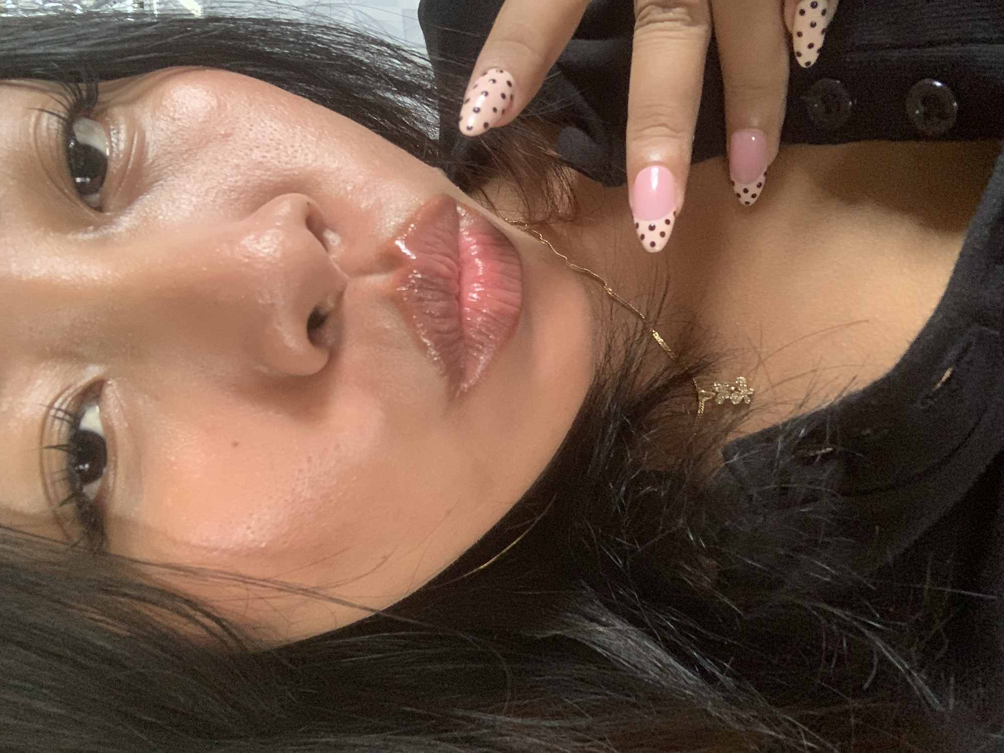 A close-up, portrait-style photograph shows a person's face and upper body, with a particular focus on their lips and hands. The person is wearing dark clothing and has long, dark hair. Their lips are coated in a glossy product, appearing plump and shiny. The person's right hand is visible, displaying manicured nails decorated with a pink base color and black polka dots. A delicate gold necklace with a charm is also visible around the person's neck. The background is out of focus, with some hints of reflective surfaces, suggesting an indoor setting. The lighting is soft and appears to highlight the dewy texture of the person's skin and lips. The image is framed tightly, cropped to emphasize facial features and cosmetic details. There is no indication of the city of Jajag, Indonesia in the image.