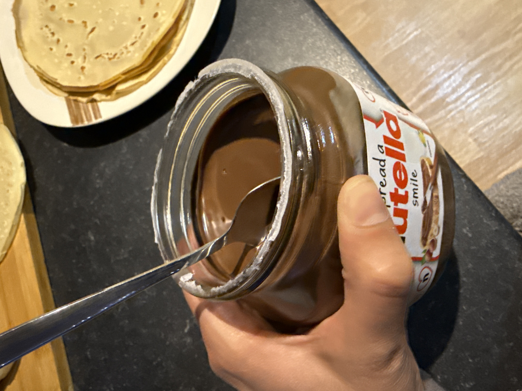 A hand holds a jar of Nutella, a spoon inside. To the side are crepes or pancakes on a plate. The location is not discernible from this image.