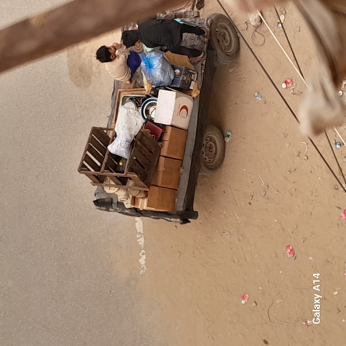 A small, overloaded vehicle sits on a dirt road. Two people are on the vehicle, one is seated, the other standing. The back of the vehicle is full of cardboard boxes, a crate, and other items. Debris is scattered on the ground nearby. A partial view of a wooden structure is visible in the foreground. The photo is taken at a high angle.