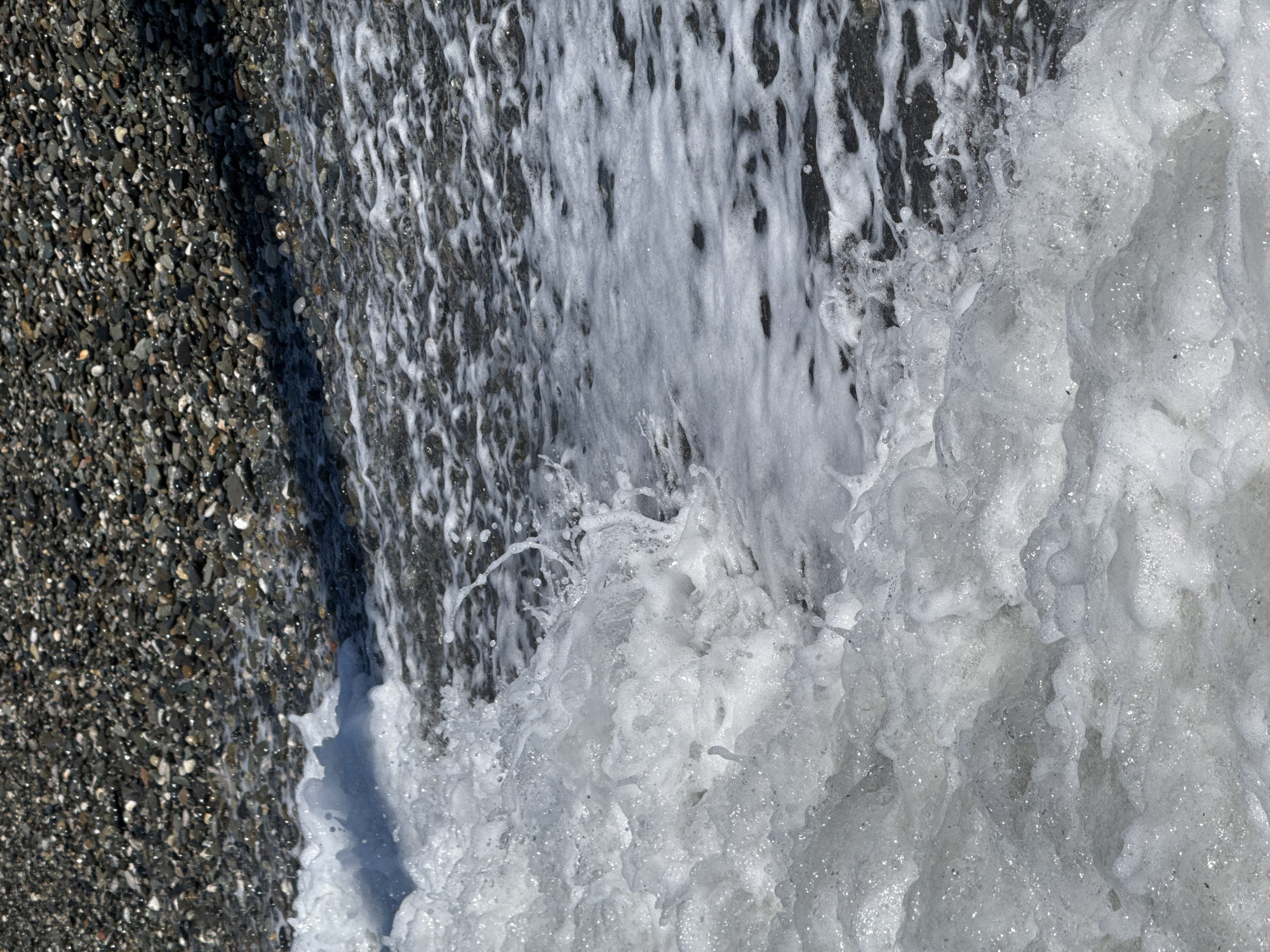 This close-up, top-down view depicts the interaction between a pebble beach and foamy seawater. The left side of the frame is dominated by a dark pebble beach, composed of small to medium-sized, predominantly grey and black stones. These pebbles appear wet and glisten with bright reflections, suggesting strong sunlight.
The right side of the image is filled with turbulent, white sea foam and water, indicative of a wave breaking or receding. The water is highly aerated, showing a myriad of bubbles and splashes, creating a dynamic and frothy texture. Streaks of white foam extend onto the darker, wet surface beneath, which could be submerged pebbles or deeper water.
The boundary between the dry-ish pebble beach and the active foamy water cuts diagonally across the image from the upper left to the lower right. The overall impression is one of natural motion and detail, captured under bright, sunny conditions. No human presence or text is visible. This scene is characteristic of a coastal environment, such as a pebble beach in Sochi, Russia.