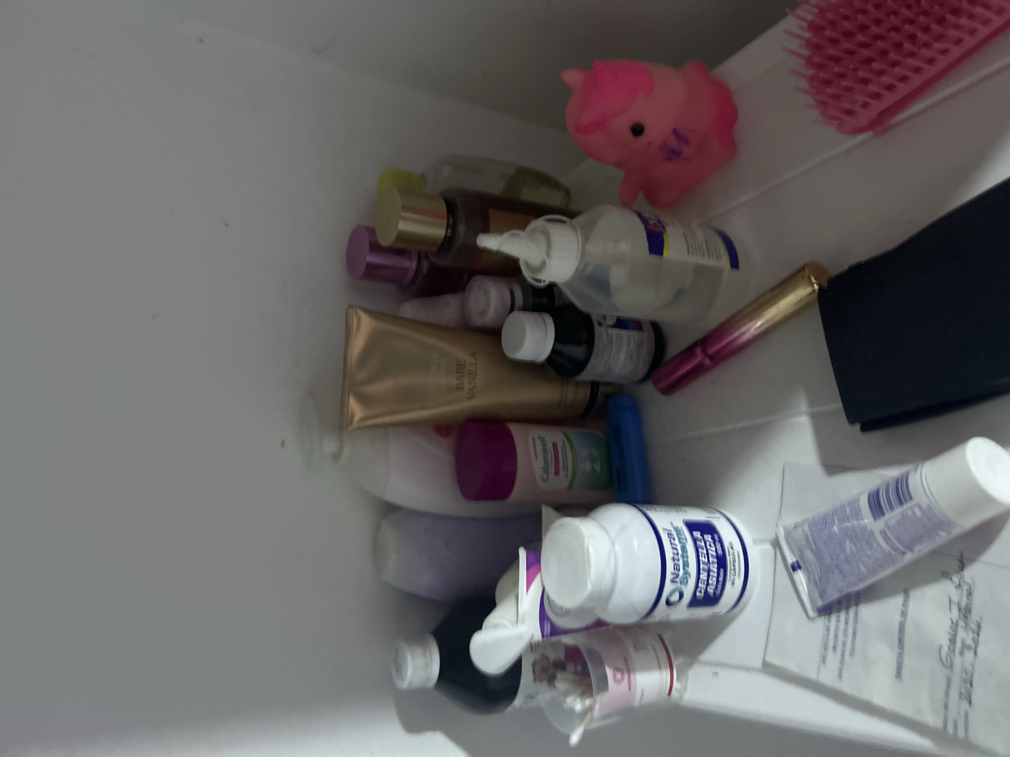 The image depicts a cluttered white shelf or container filled with various items. The items include: a pink toy pig, a pink hairbrush, multiple bottles of liquids (some with white labels and blue text, some clear, and some dark), a tube of gold-colored lotion labeled "Bare Vanilla," a tube of purple cream, a black rectangular object, and a tube of blue and white cream. There are also documents. The scene is not explicitly identifiable as being located in San Juan de Los Morros, Venezuela. The environment appears confined. There are no people visible. The image does not reveal any actions or interactions.