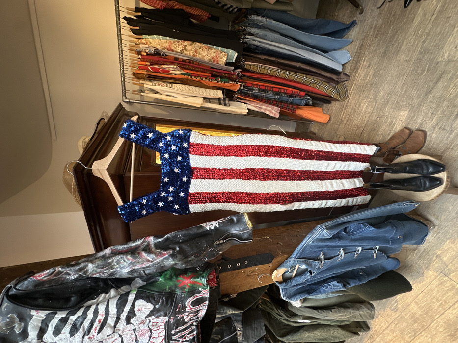 This image depicts the interior of a vintage clothing store located in Paris, France. The central subject is a sleeveless, form-fitting dress adorned with sequins, designed in the pattern of the American flag. The upper portion of the dress is blue with white stars, while the lower body consists of alternating vertical red and white stripes. It hangs on a wooden hanger from the top of a dark wooden armoire.
To the left of the dress, a black leather jacket, heavily embellished with white and red painted designs, hangs from a wooden support. Below this jacket, a blue denim jacket with silver metal toggle closures is displayed on a wooden hanger, positioned above other military-style olive-green garments. A black belt hangs vertically from a support near these jackets.
On the right side of the scene, a metal clothing rack is densely filled with a variety of garments, including patterned shirts, trousers, and jeans. On the light-colored wooden floor directly beneath the sequined dress, two pairs of ankle boots are arranged on a small, round wooden stool: a pair of black pointed-toe boots in the foreground and a pair of brown square-toe boots behind them. The store's interior features light-colored walls and a light wooden floor. Several straw hats are visible on top of the dark wooden armoire.