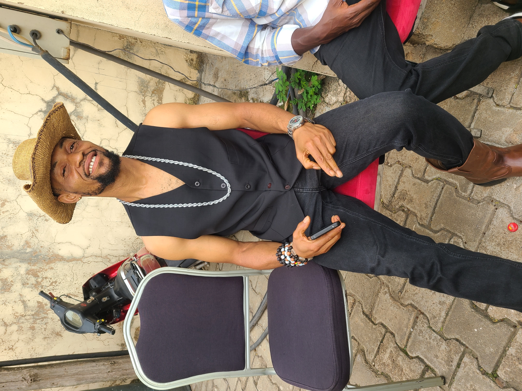 The image depicts a man wearing a straw hat, a black vest, and black jeans, seated on a black chair in what appears to be an outdoor setting. He is holding a black mobile phone in his right hand and wearing a silver watch on his left wrist. Multiple beaded bracelets are also visible on his left wrist. Behind him, a red motorcycle is parked. To his right, another person, partially visible, is wearing a blue and white plaid shirt and black trousers. The ground is paved with interlocking concrete blocks. The background features a textured, light-colored wall with some visible wiring and conduit. A small green plant is growing near the wall. The scene is likely located in Barakin Lerawi, Nigeria, given the context of the query.
