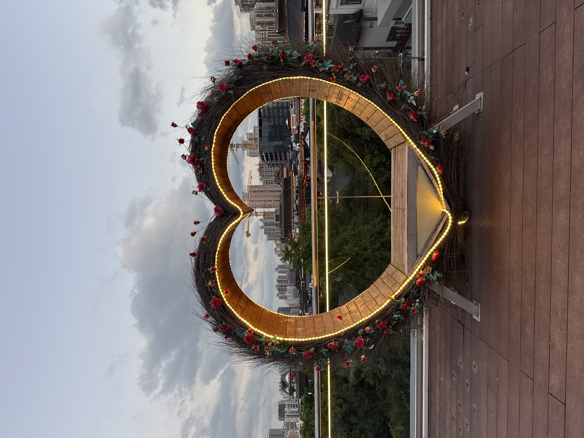 The image shows a large, heart-shaped decoration made of twigs and adorned with red roses and warm-toned lights. It is positioned on a wooden deck, overlooking a cityscape at dusk. In the background, various buildings, including residential towers and traditional Chinese architecture, are visible. Construction cranes suggest ongoing development in the area. The sky is overcast with diffused light. The location is a rooftop, likely in Shanghai, China, given the architectural styles and the presence of construction cranes. There are no people or animals present in the image.