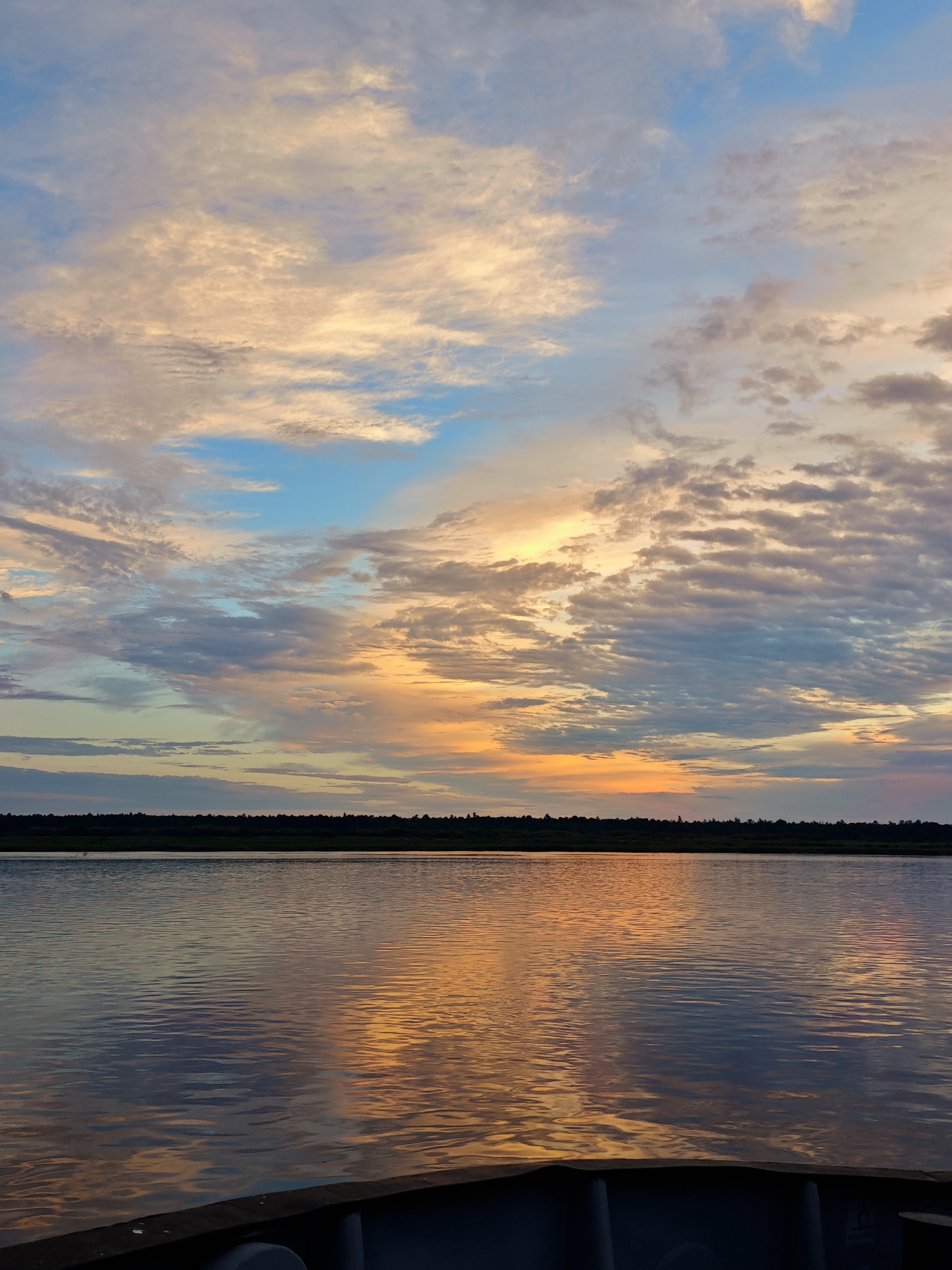 This is a photograph of a sunset over a body of water, viewed from what appears to be the front of a boat. The sky is filled with a variety of clouds, some white and wispy, others darker and more textured. The clouds are illuminated by the setting sun, creating a gradient of colors from warm oranges and yellows near the horizon to cooler blues and purples higher up.
The water in the foreground is calm, with gentle ripples reflecting the colors of the sky. A dark, indistinct object, possibly the edge of the boat, occupies the very bottom of the frame. In the distance, a dark line of trees and vegetation forms the horizon, separating the water from the sky. The overall mood of the image is peaceful and serene, evoking the beauty of a natural landscape at dusk. The time of day is clearly sunset. There is no visible text in the image.