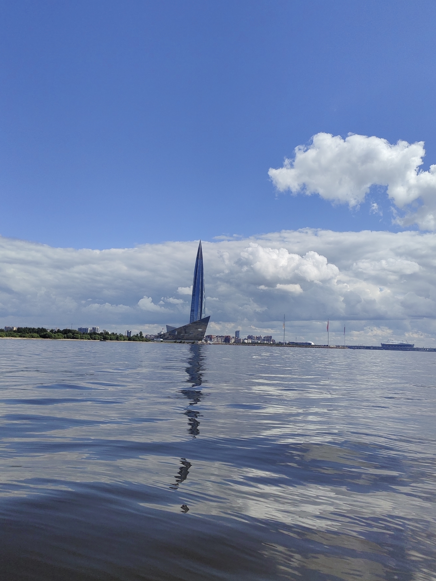 The image captures a view of the Neva River in Saint Petersburg, Russia, on a clear day with scattered clouds. Dominating the scene is the Lakhta Center, a striking modern skyscraper with a distinctive sail-like design, reflecting the sky and surrounding water. The skyscraper is located on the outskirts of the city, to the left of the image, with a low-rise cityscape and a green shoreline stretching behind it. The foreground of the image is filled with the rippling surface of the river, showing reflections of the sky and the distant structures. The overall impression is one of urban development integrated with natural water bodies under a bright, blue sky.