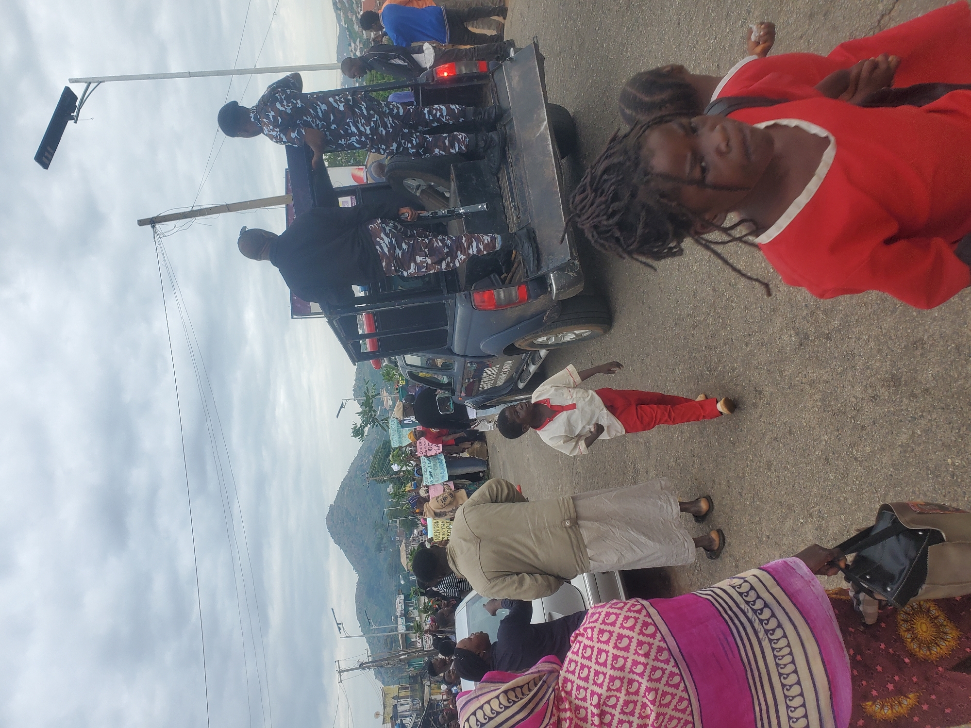 The image depicts a street scene in Gwong, Nigeria, under a cloudy sky. A group of people, including adults and children, are gathered on a paved road. In the foreground, a young child in a red shirt and pants walks away from the camera. To their left, a woman in a pink and white patterned outfit carries a black handbag. Behind her, a man in a beige robe and sandals stands near a silver car.
Further back, a dark-colored pickup truck, possibly a police vehicle due to the presence of individuals in camouflage uniforms, is parked. Two individuals in camouflage uniforms are visible in the back of the truck, one holding what appears to be a rifle. Other people are seen standing around the truck and in the background. Some individuals are holding signs, suggesting a protest or demonstration. Beyond the immediate scene, buildings and a hilly landscape with vegetation are visible. Power lines and streetlights are also present. The overall atmosphere appears to be one of public gathering and possibly civic action.