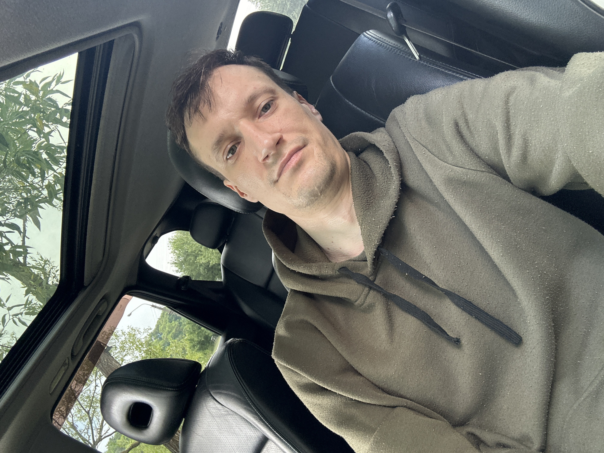 A man is taking a selfie inside a car with a sunroof open. The man is wearing a green hoodie and is seated in the driver's seat. The interior of the car is visible, including the dashboard and the passenger seat. Outside the car, trees and foliage are visible, suggesting a natural or semi-rural setting. The image does not contain any specific geographical markers that would indicate the location is Makiv, Ukraine.