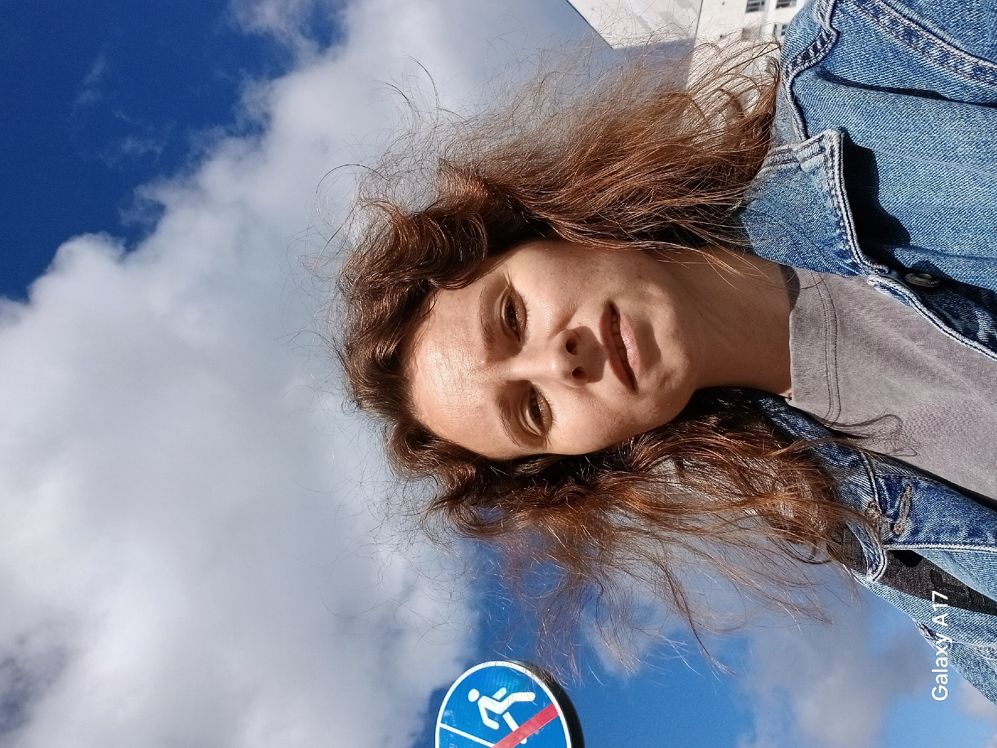 An outdoor, low-angle, close-up selfie shows a woman with reddish-brown, windblown hair against a bright blue sky with white clouds. She is wearing a denim jacket over a grey t-shirt. To the left of the woman is a blue traffic sign with a white outline depicting a pedestrian walking with a red diagonal line crossing it, indicating "No Pedestrian Crossing." A building is visible on the far right of the image. The text "Galaxy A17" is in the bottom left corner. The scene appears to be in a city environment, possibly Yekaterinburg, Russia, given the presence of the traffic sign and urban architecture.