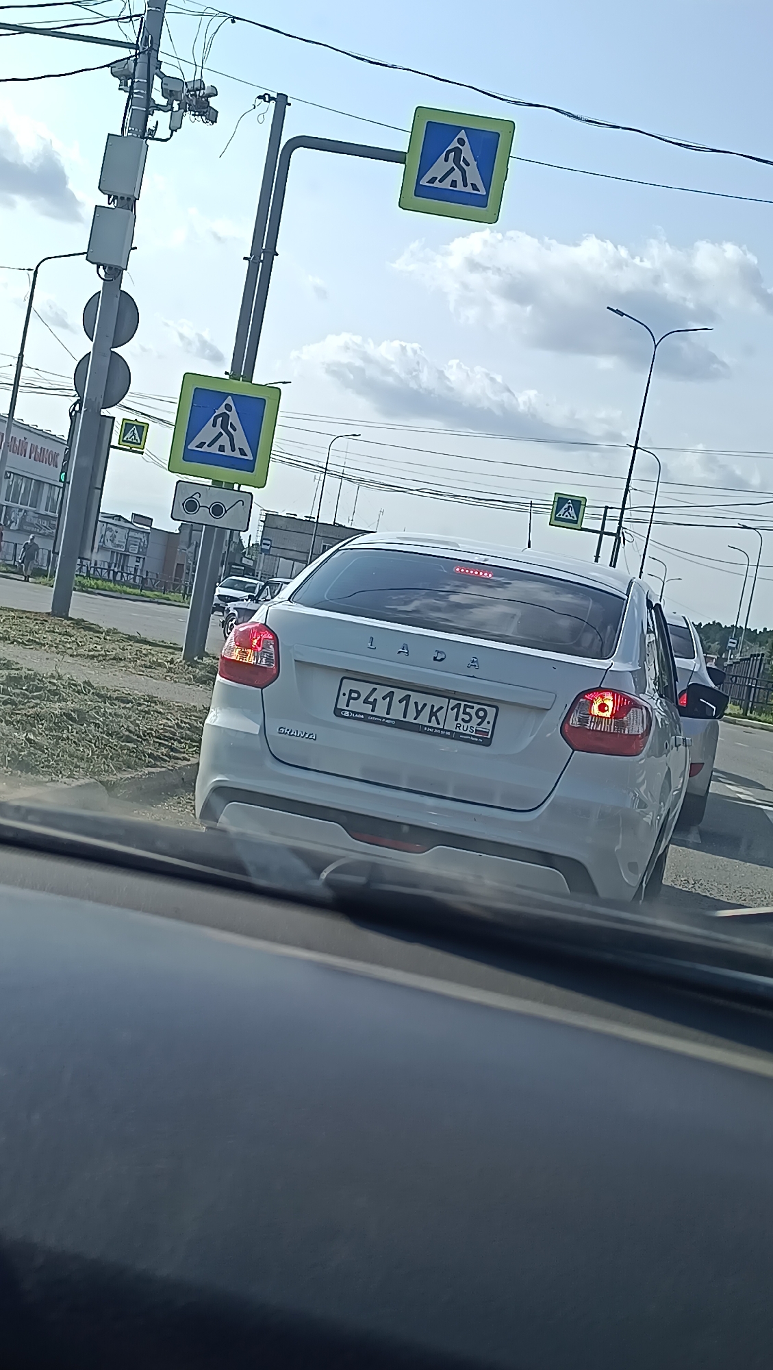 The image shows a view from inside a vehicle looking out at traffic and a street scene. In the foreground, the dashboard of the vehicle is visible. In the middle ground, a white Lada car is stopped at what appears to be an intersection. The license plate of the Lada reads "P411YK 159". Pedestrian crossing signs are visible on poles on the right side of the road. In the background, there are buildings, streetlights, and power lines. The sky is partly cloudy. The scene is consistent with an urban or suburban environment. The image appears to have been taken in the city of Chaikovsky, Russia, based on the presence of Russian license plates and street signs.