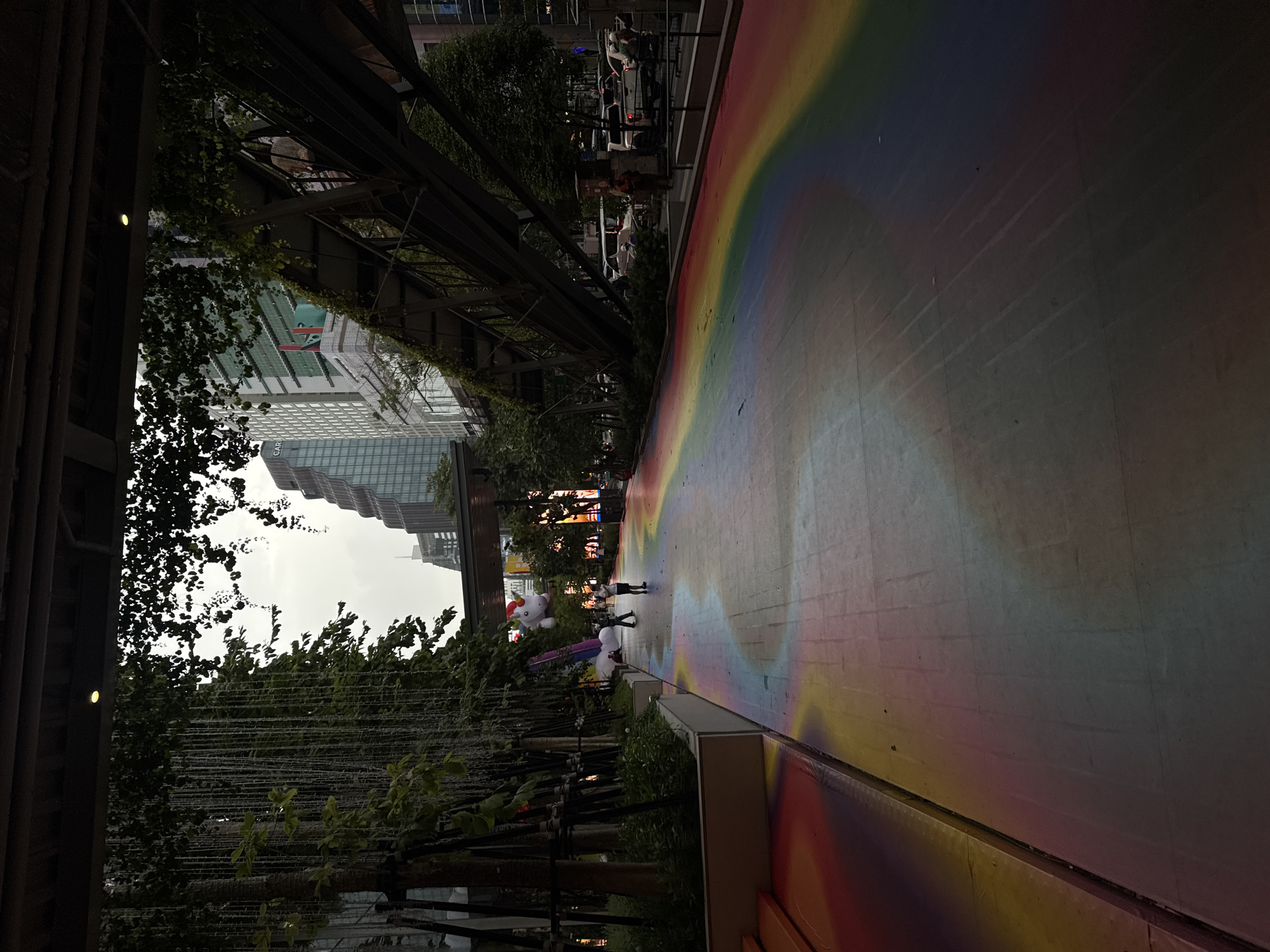 The image captures a paved walkway in a city, likely Bangkok, Thailand, adorned with a vibrant rainbow-colored design. Tall buildings, some with reflective glass facades, dominate the background. Lush green trees and hanging foliage frame the scene, suggesting a well-landscaped urban area. A pedestrian overpass or bridge with metal structures and overgrowth crosses the upper right portion of the frame. In the foreground, a few individuals are present on the walkway, one appearing to be a child. A large inflatable unicorn can be seen on the left side of the walkway, suspended in the air. The overall atmosphere is one of a modern, bustling cityscape with touches of nature and artistic expression.