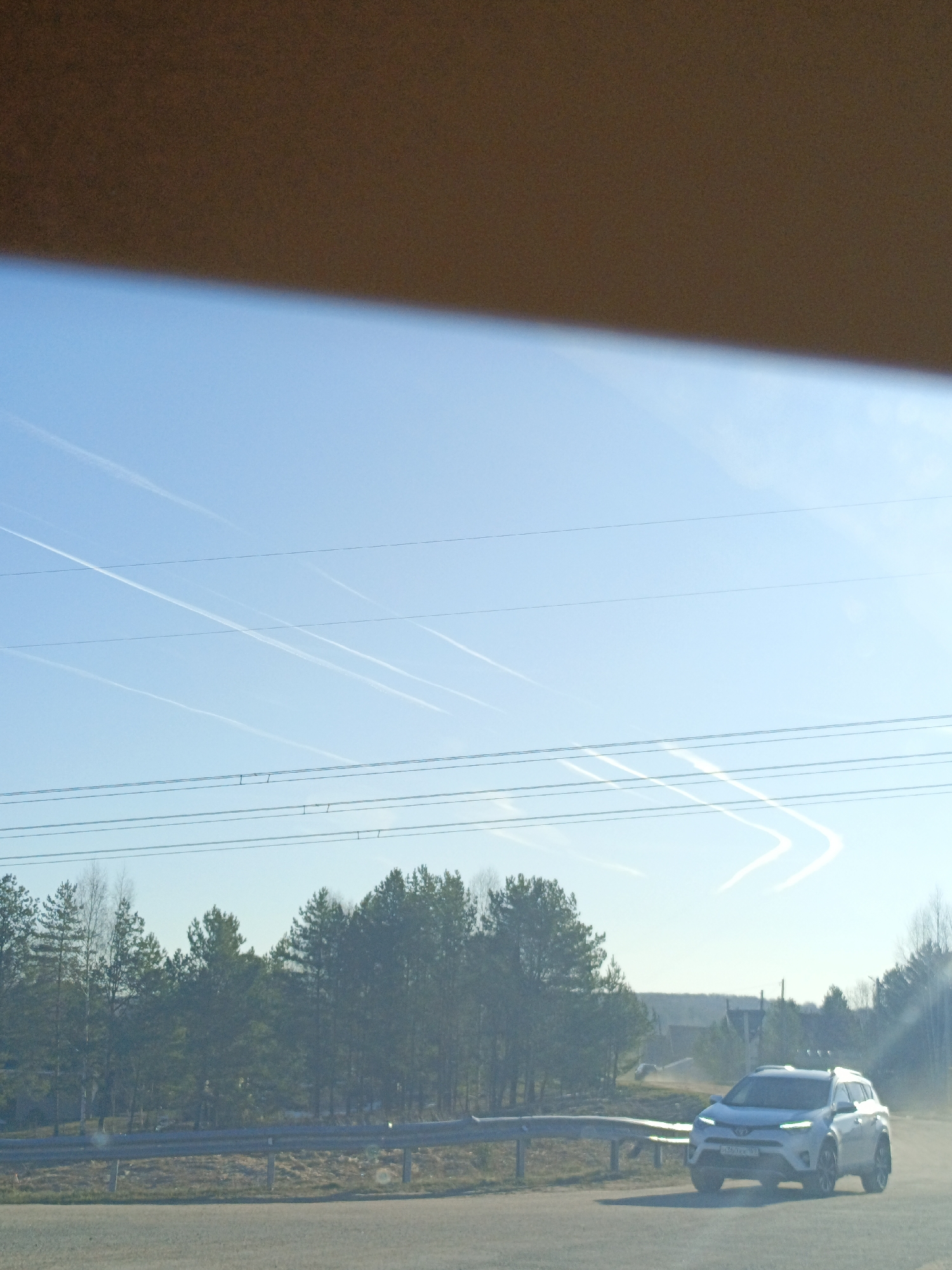 The image shows a white SUV on a paved road, with trees in the background and a blue sky with contrails. Power lines are visible in the sky. Based on visual analysis, the location is likely Dubovaya, Russia. The photo appears to be taken from inside a vehicle.