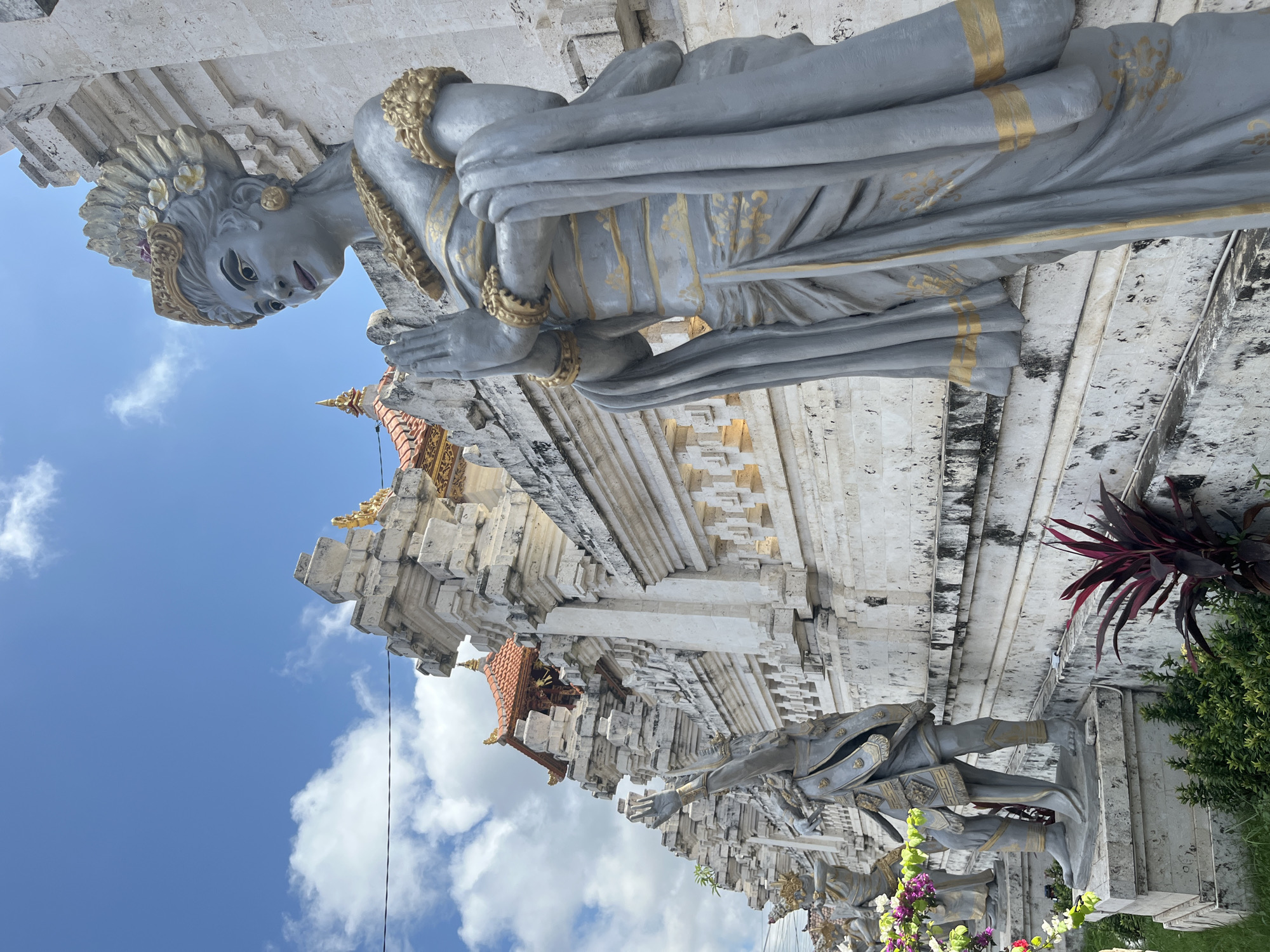 The image shows a row of gray stone statues in front of a white stone wall at a temple in Kuta Selatan, Indonesia. The statue in the foreground is a woman with her hands clasped in prayer, adorned with gold accents on her headdress, jewelry, and clothing. Behind her, to the left, is another statue of a man with one arm raised. The wall behind the statues is intricately carved and topped with a traditional Balinese tiled roof. The sky is blue with scattered white clouds. Green foliage and some colorful flowers are visible in the lower left corner of the frame.