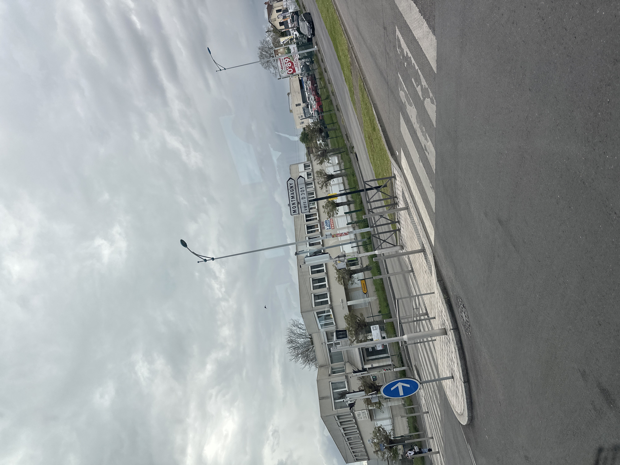 The image shows a street scene in Sarcelles, France. The view is taken from a moving vehicle, looking towards the side of the road. A multi-story, light grey building with repetitive window patterns runs along the road. Various street signs are visible, including directional signs (e.g., a blue sign with an upward-pointing arrow) and a sign indicating "Montmagny vers D 301". Other signs and commercial advertisements are present further down the road. A crosswalk is seen painted on the road. Street lamps are positioned along the sidewalk. The sky is overcast with a pattern of cloud cover. No people are readily visible.