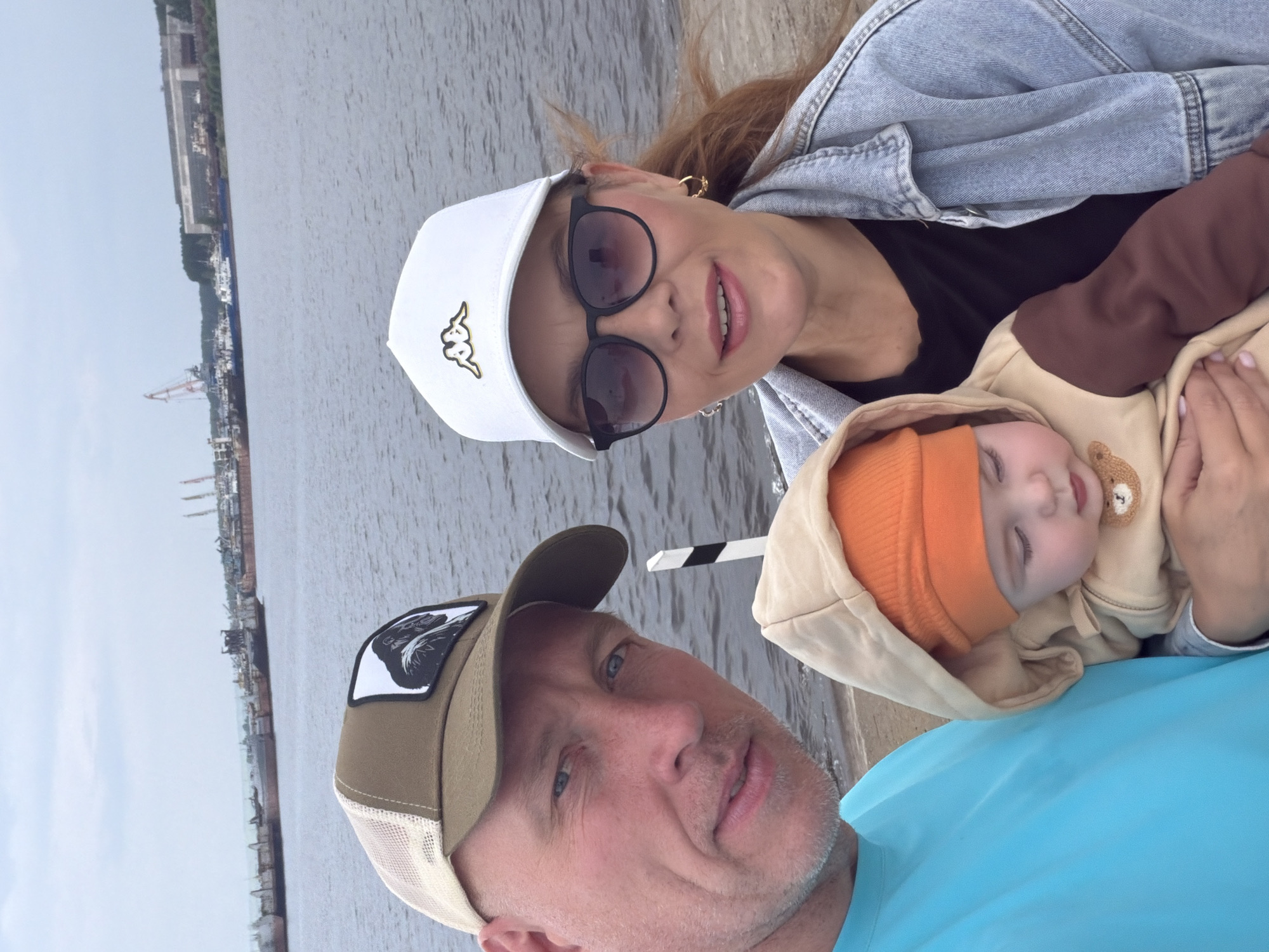 This is a selfie-style photo of three people. A man in the foreground is wearing a tan baseball cap with a patch featuring a black and white bird. He has a light complexion and is looking towards the camera with a slight smile. Behind him, a woman is wearing a white baseball cap with a gold logo, sunglasses, and a denim jacket. She is smiling at the camera and holding a baby. The baby is wearing an orange beanie and a tan hooded jacket with a bear detail. The baby appears to be sleeping. The background shows a body of water, possibly a river or lake, with a long industrial shoreline featuring cranes and buildings. The sky is overcast, suggesting a cloudy day. The overall mood of the photo is casual and familial. The image appears to have been taken outdoors during daylight.