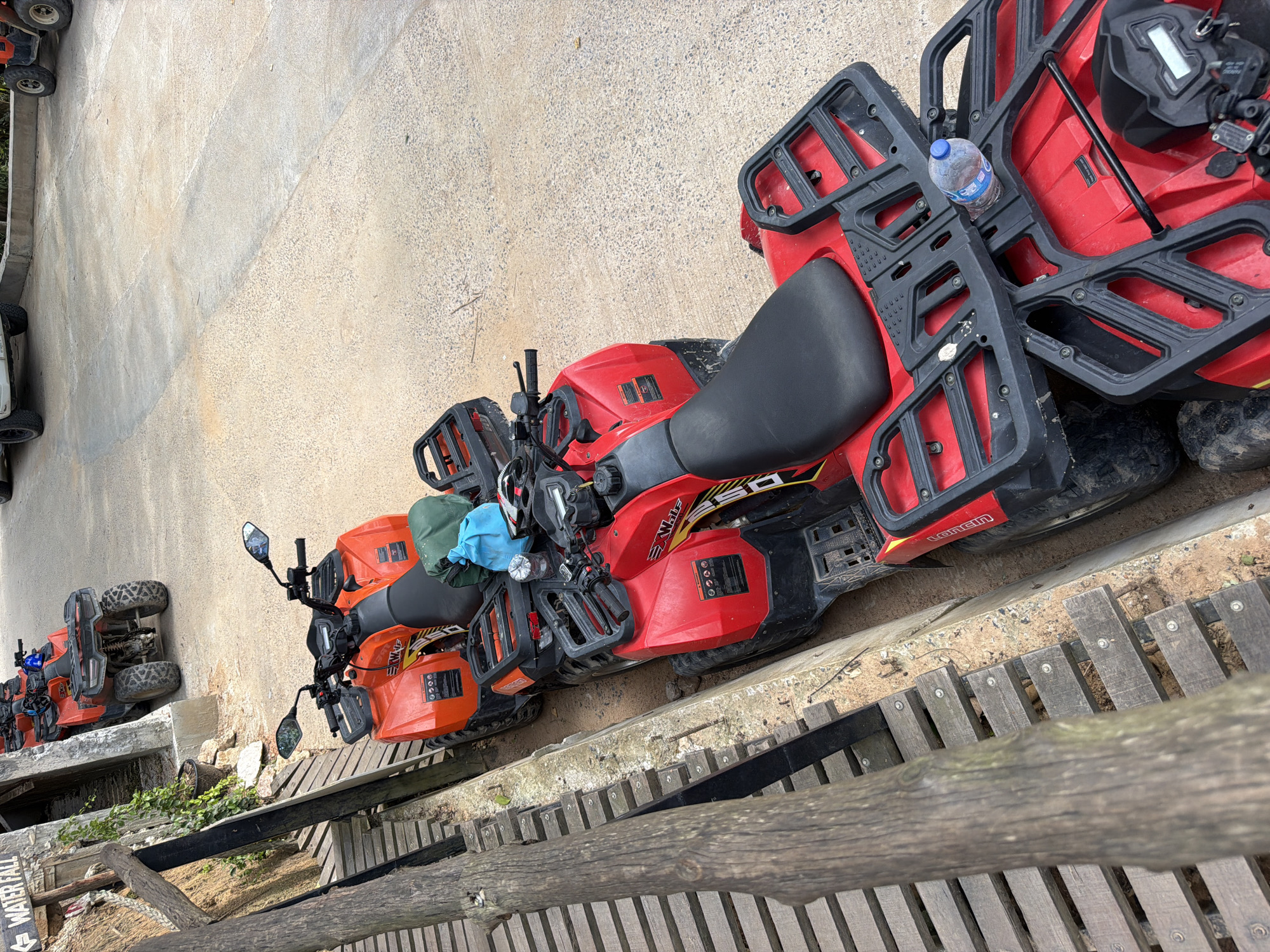 The image depicts an outdoor scene at an ATV rental facility, possibly near a waterfall, in Ko Samui, Thailand. Multiple red and orange ATVs are parked in a line on a concrete surface next to a wooden walkway. The ATVs are equipped with racks and some have bags and a water bottle on them. The walkway has a sign partially visible that says "WATER FALL". The ground is a mixture of sand and gravel. The environment suggests a tourist attraction offering adventure activities. There are no people visible in the image.
