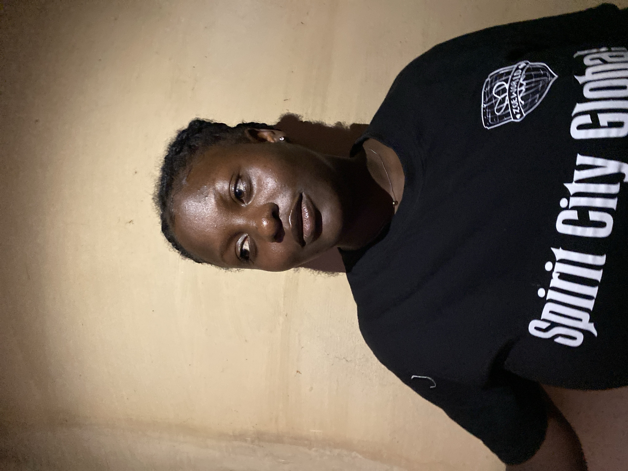 A young Black woman is pictured indoors against a tan-colored wall. She is wearing a black t-shirt with white lettering reading "Spirit City Global," and a logo above it. The image was likely taken in Kuru Dungus, Nigeria.