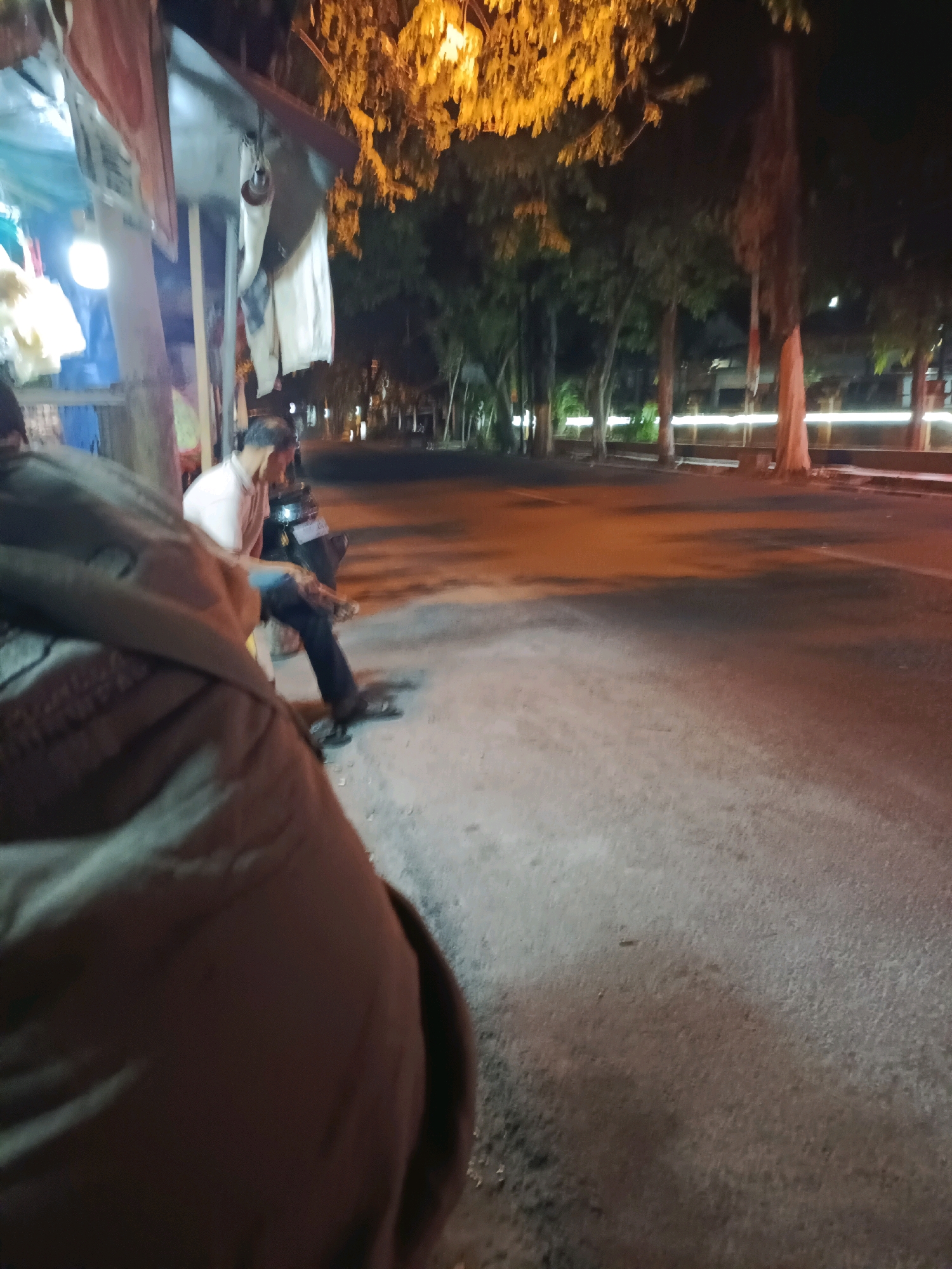 A blurred, nighttime photograph taken in Lamongan, Indonesia, captures a street scene with several individuals and elements of an urban environment. In the foreground, the back of a person wearing a brown jacket is visible, partially obscuring the view. To the left, a stall or small building is apparent, with tarps, white cloth, and a light fixture hanging. A man in a light-colored shirt and dark pants is seated on a motorcycle, appearing to be engaged in conversation or waiting. The background reveals a street with trees lining its edge, illuminated by streetlights and ambient light from nearby buildings. The overall impression is that of a casual evening gathering or stop along a roadside in an Indonesian city.