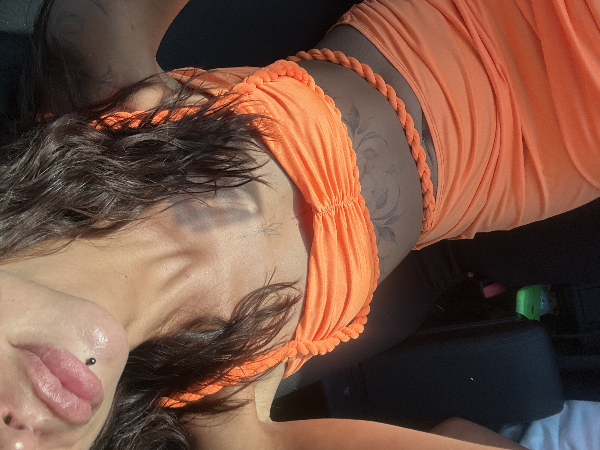 A young woman with dark hair and a piercing on her lower lip is wearing a ruched, orange-toned bikini top with braided straps and a matching skirt. She has a tattoo on her abdomen. She is seated in a vehicle, with the interior visible behind her. The scene is illuminated by natural sunlight. The location is not specified in the provided image or context.
