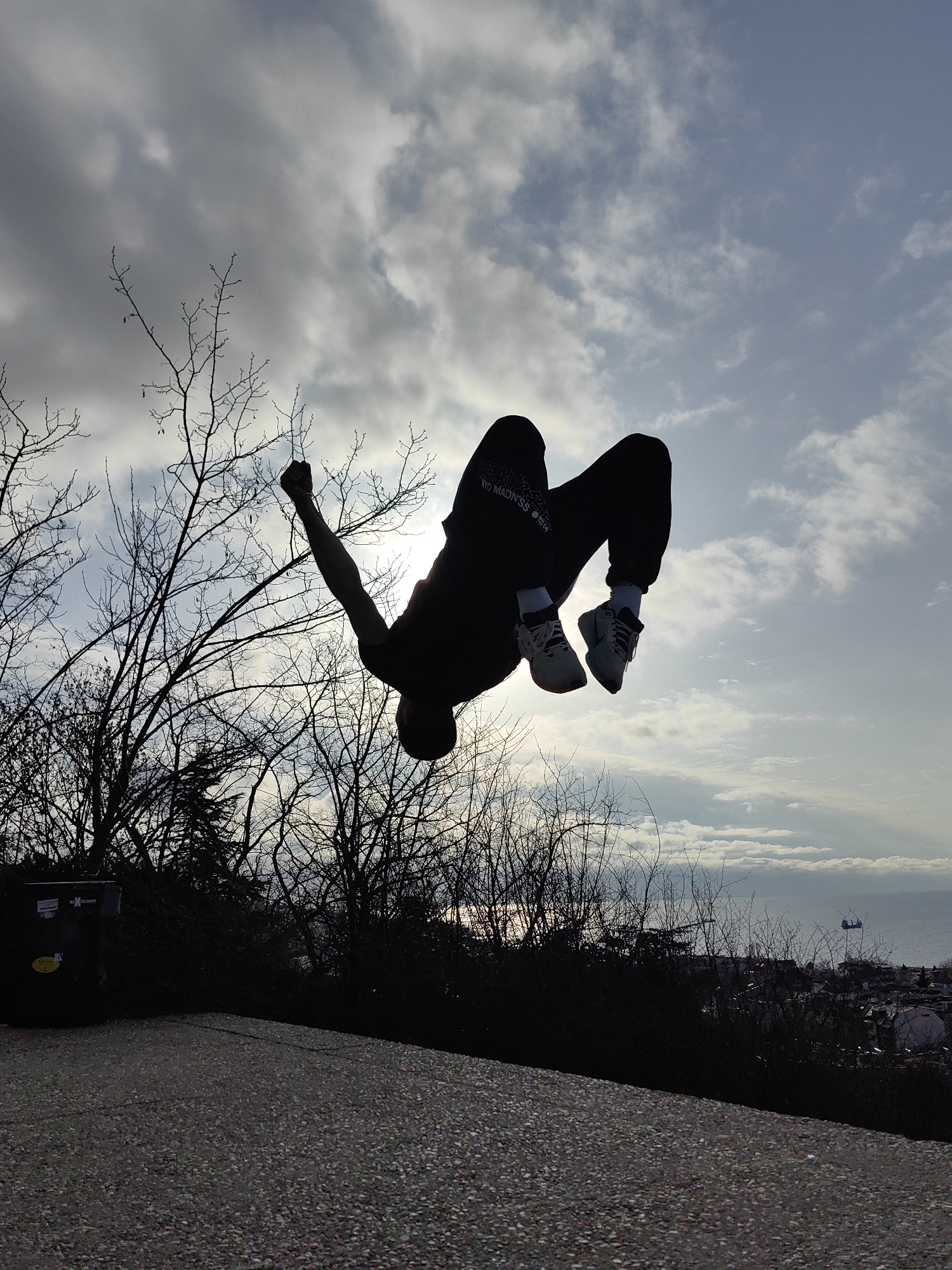 A silhouetted figure performs a backflip against a cloudy sky. The figure is wearing dark pants and light-colored shoes. Branches from trees extend into the frame on the left side. A trash can is visible near the bottom left. In the distance, the Black Sea and the city of Varna, Bulgaria, can be seen. The ground is paved.