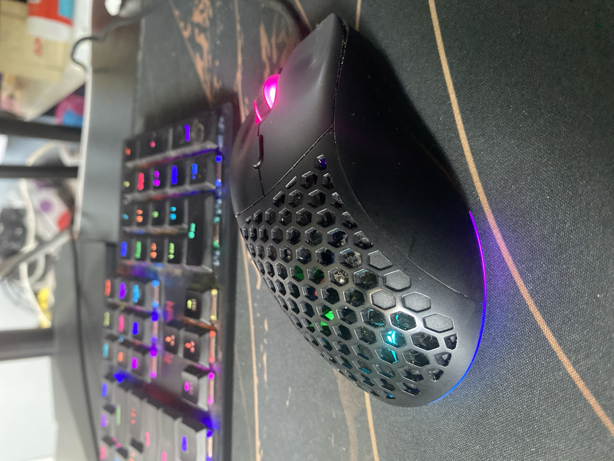 This is a close-up, eye-level shot of a black, honeycomb-patterned gaming mouse with RGB lighting, placed on a black mousepad with subtle brown lines. The mouse has a pink illuminated scroll wheel and a light blue and purple glow emanating from its base. In the blurred background, a portion of a colorful, RGB-lit keyboard is visible. There are no people or animals in the image. The scene appears to be an indoor setting, likely a desk setup. The location in the city of Kozet, Russia, is not discernible from the image itself.