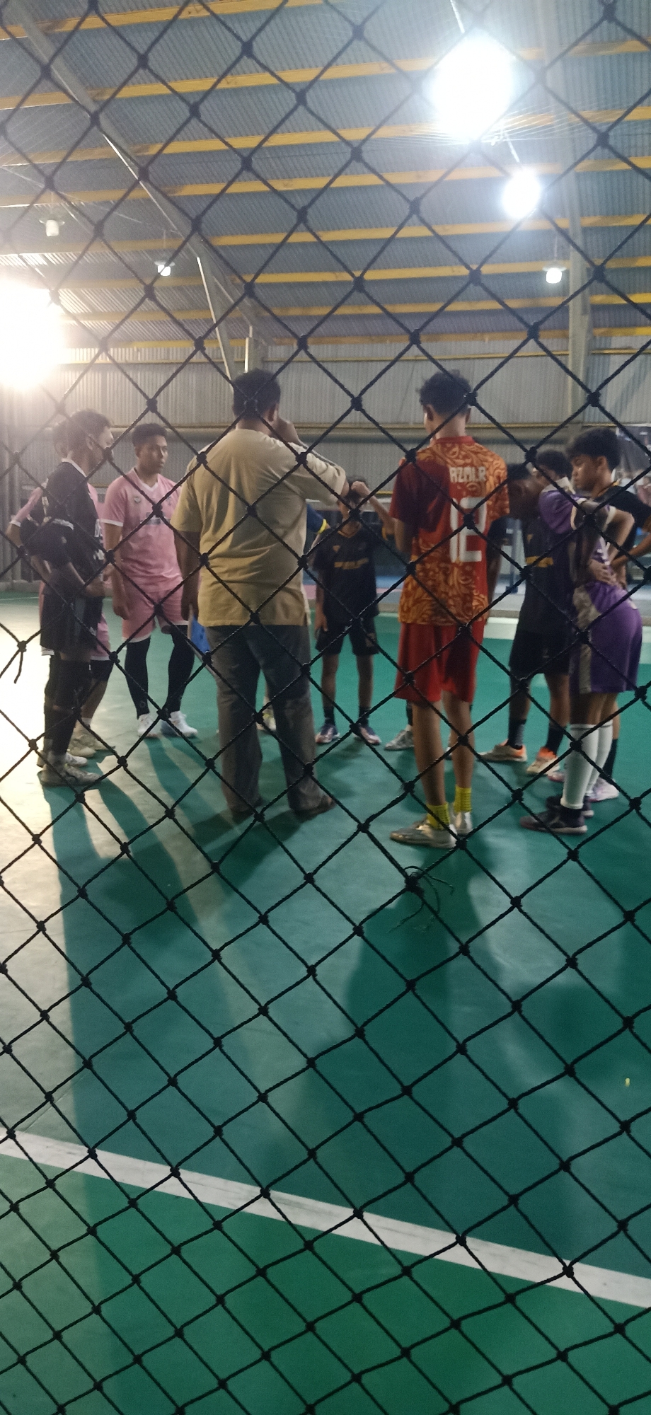 A group of young men, likely a soccer team, are gathered in a huddle on a green indoor sports court. They are wearing a variety of athletic attire, including jerseys, shorts, and cleats. The court is lit by overhead fluorescent lights, and the scene is viewed through a black mesh net. The background shows the industrial-style ceiling of the indoor facility, with corrugated metal and yellow beams. The overall impression is one of a casual gathering after a practice or game, possibly at the Palohgadeng indoor soccer field in Indonesia, where such facilities are common.
