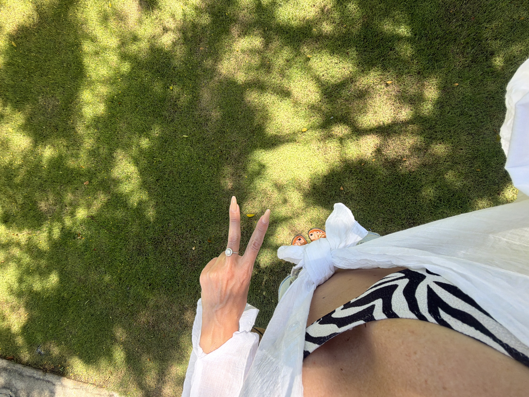 In a close-up, top-down shot, a person's hand wearing a ring with a large diamond is positioned to create a peace sign. The hand is placed over a zebra-print bikini top, which is partially covered by a white sheer garment. Orange-soled sandals are visible beneath the white garment. The background consists of lush green grass dappled with sunlight filtering through tree leaves, creating a shadow pattern. A portion of a gray concrete path is visible in the upper left corner. The scene appears to be an outdoor setting, likely a garden or park in Dong Ninh Hoa Ward, Vietnam.