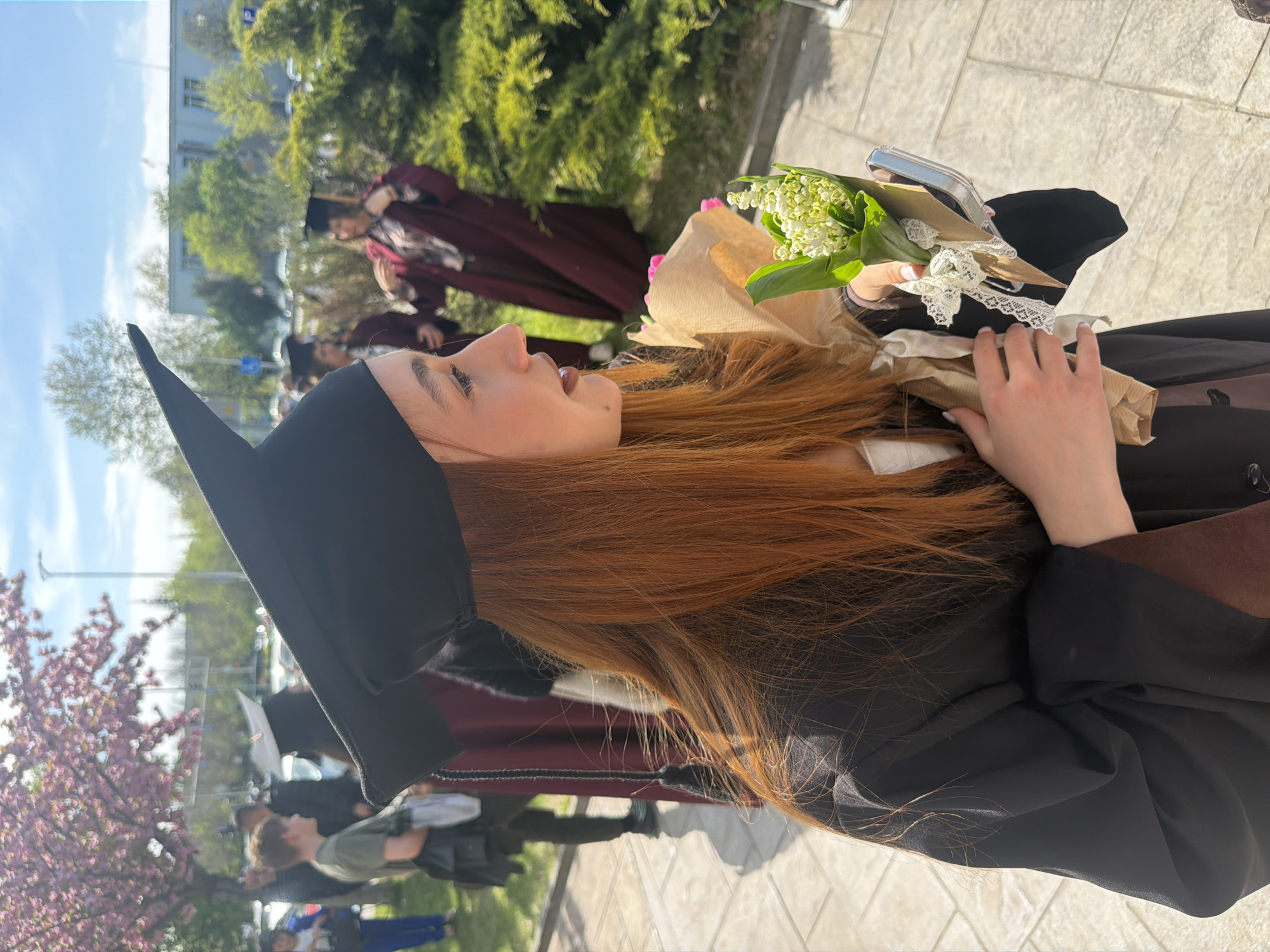 A young woman is captured in profile, looking upwards and slightly to her right, under a bright, sunny sky. She has long, flowing reddish-brown hair and is wearing a black graduation cap with a pointed front and a black graduation gown, possibly over a maroon or brown garment. In her hands, she holds a small bouquet of white flowers, resembling lily of the valley, wrapped in brown paper and tied with white lace. A silver object, possibly a mobile phone, is also visible near the bouquet. Her mouth is slightly open, suggesting she might be speaking or smiling.
The background is softly out of focus, revealing several other individuals, some of whom are also dressed in academic robes, primarily maroon, hinting at a graduation or similar academic event. Lush green trees, including some with vibrant pink blossoms, frame the scene, suggesting springtime. A light-colored building with visible windows and a paved ground area can be seen in the distance. The setting appears to be outdoors during the daytime in Plovdiv, Bulgaria. No discernible text is legible within the image.